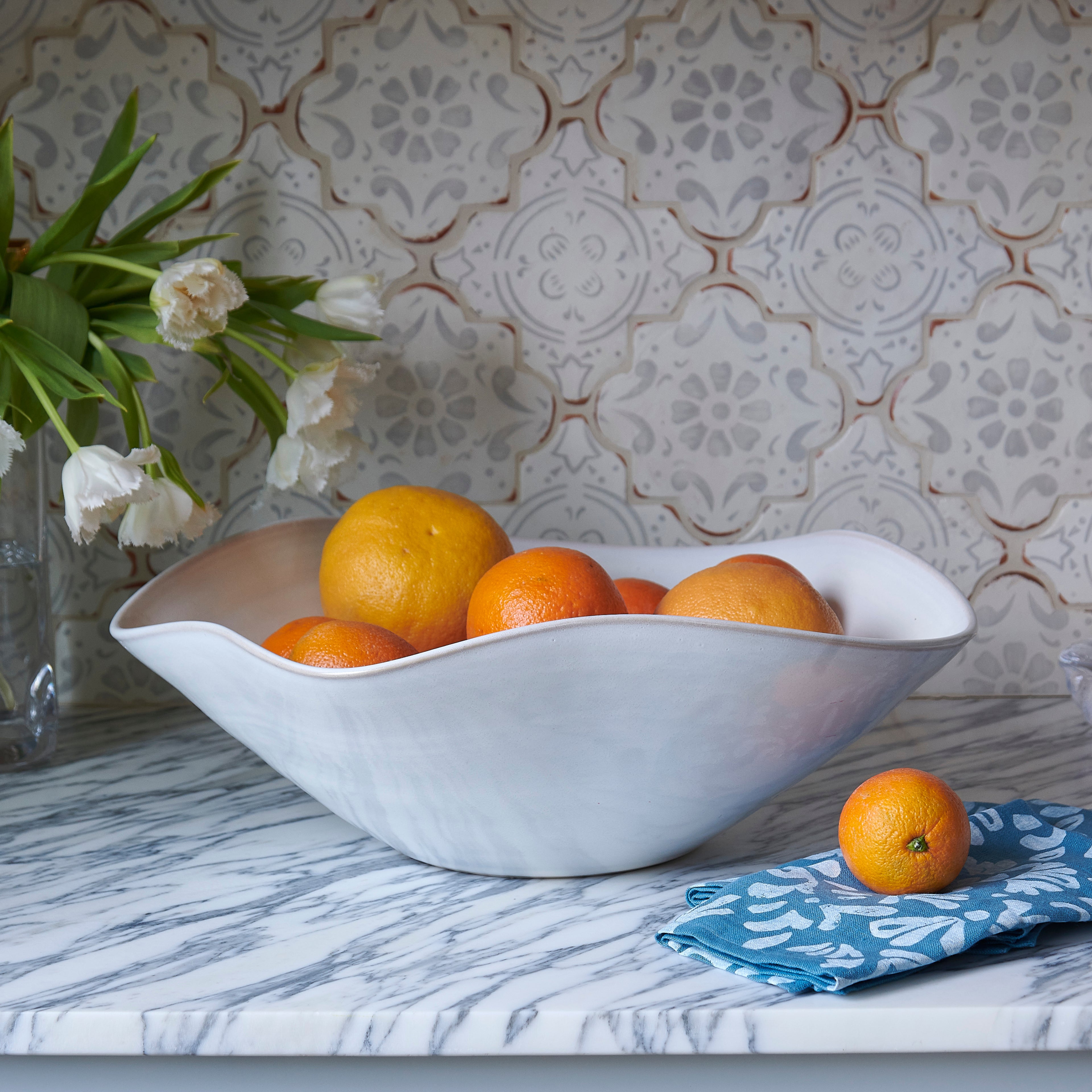 White ceramic bowl with wavy edges holding oranges and lemons, displayed on marble surface.