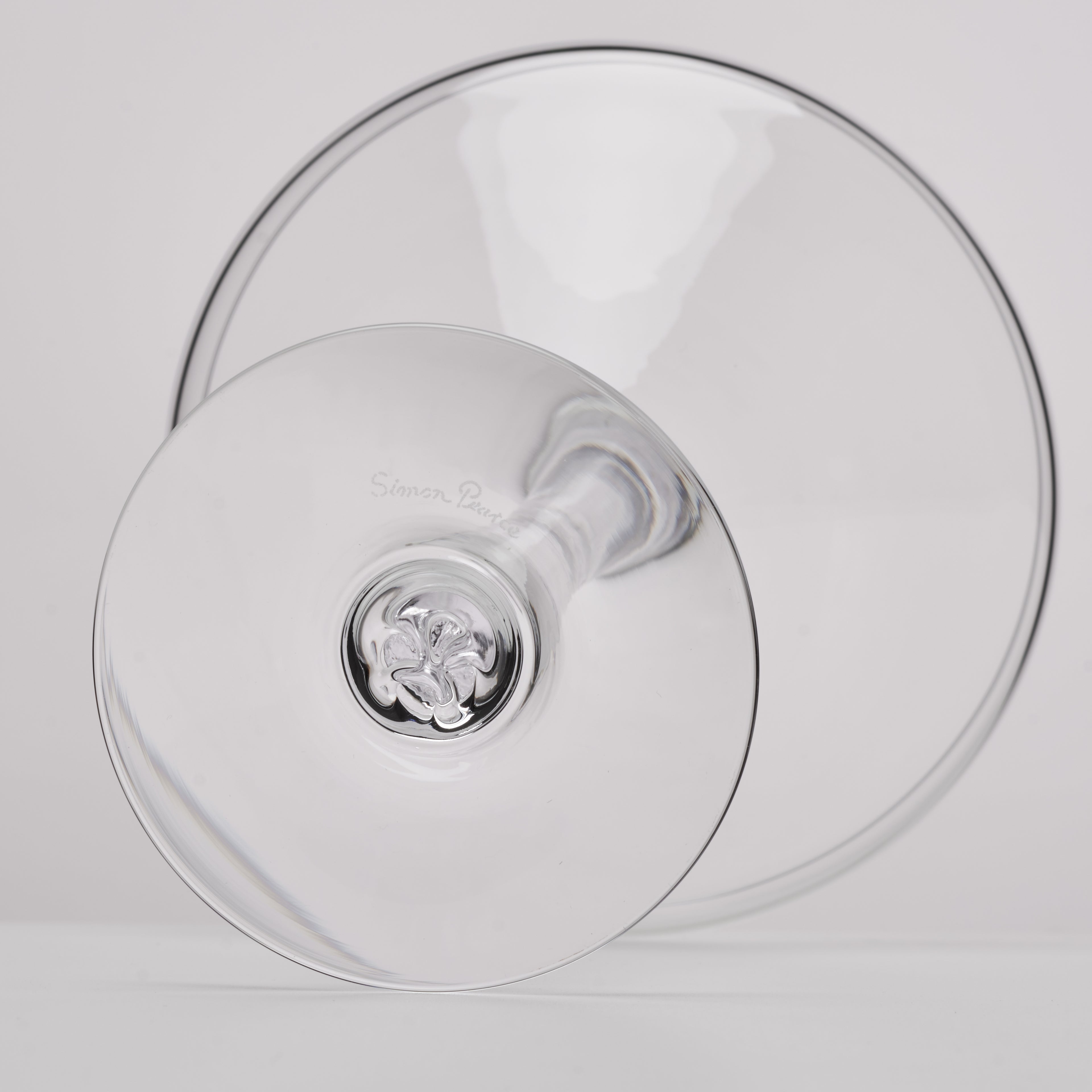 A clear glass wine glass with a visible stem and circular base, photographed on a clean light gray background. A faint engraved brand name appears on the glass.