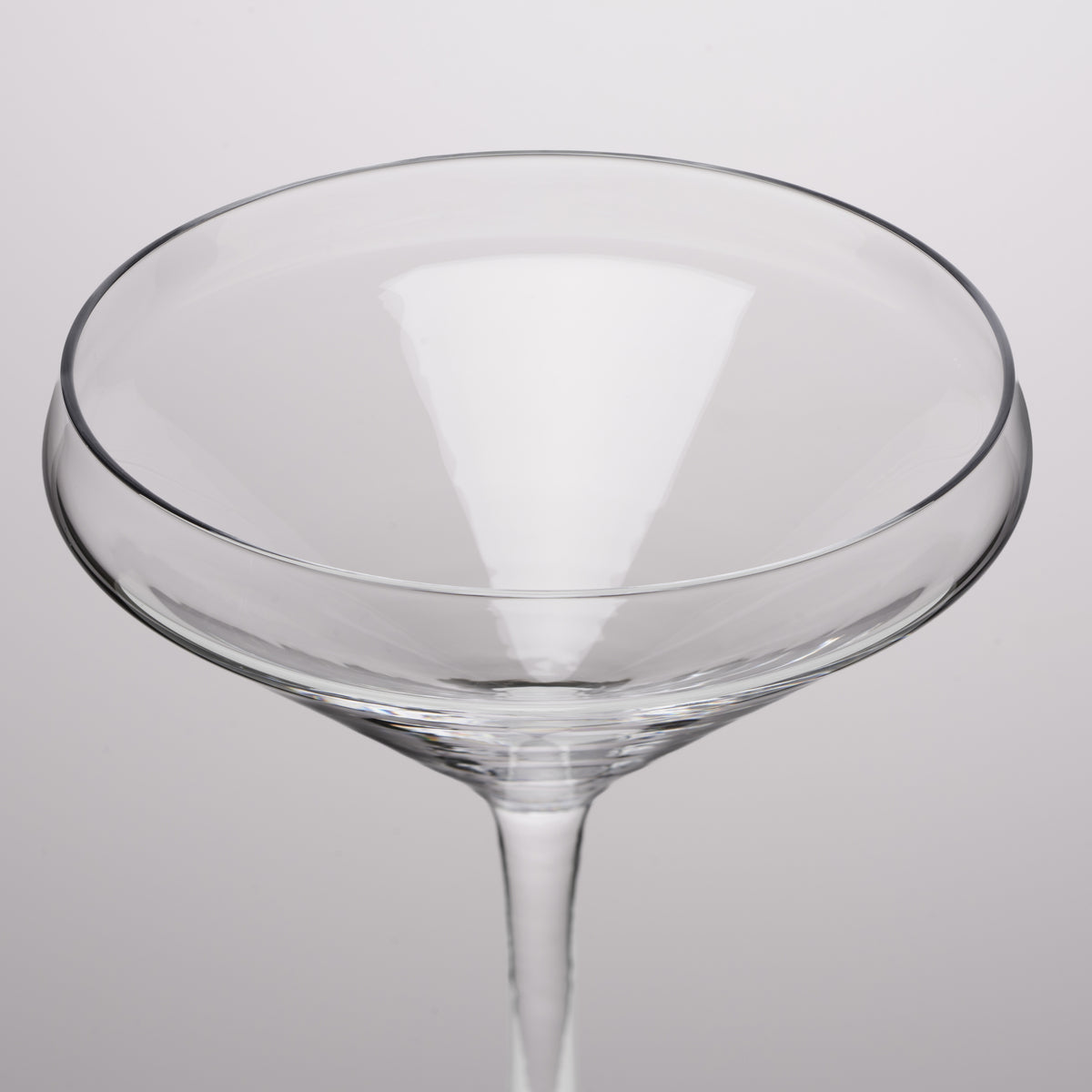 A clear glass coupe glass with a wide, shallow bowl and a slender stem, shown against a neutral light gray background.