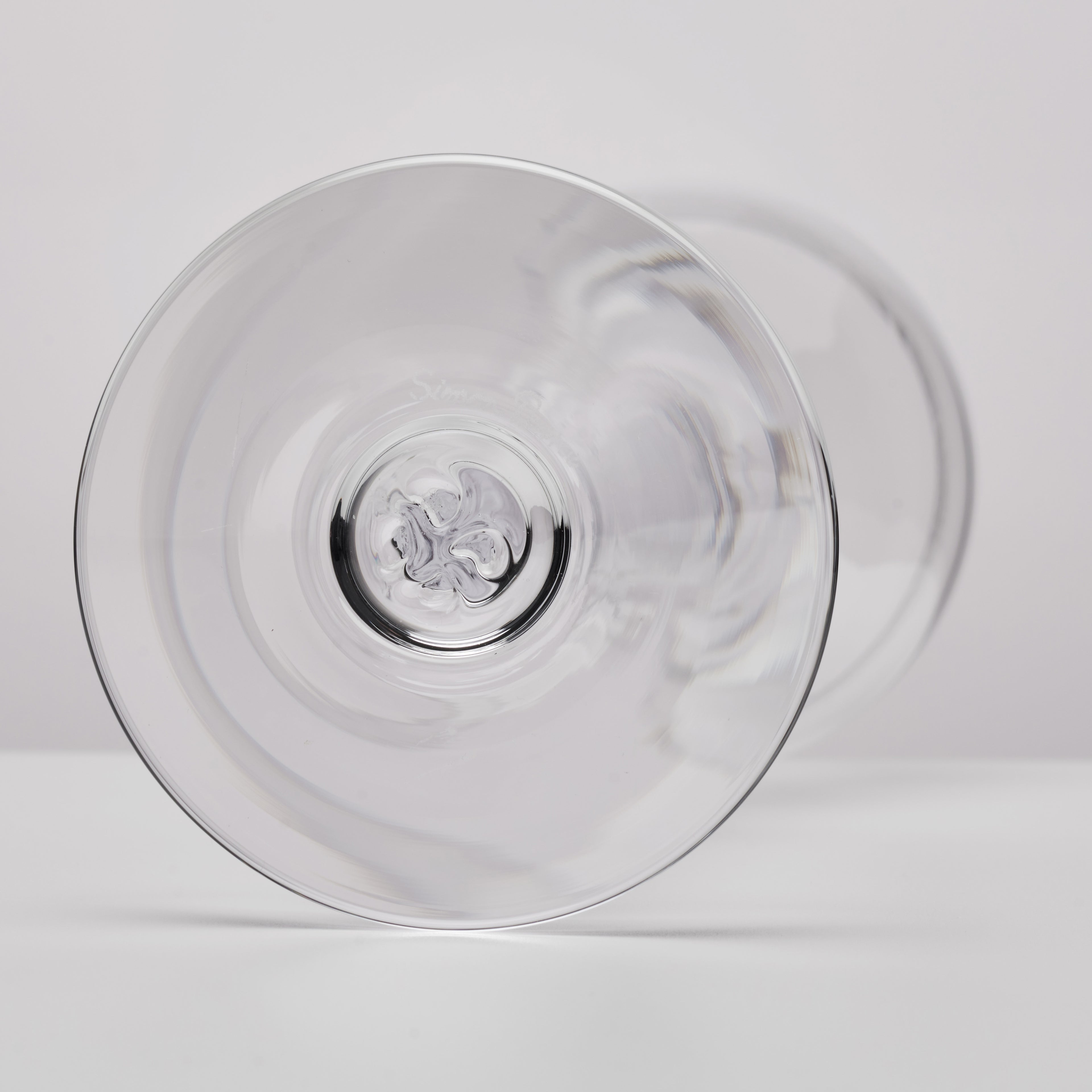 A clear glass coupe glass shown from a bottom angle, highlighting its circular bowl and stem base, set against a clean white background.