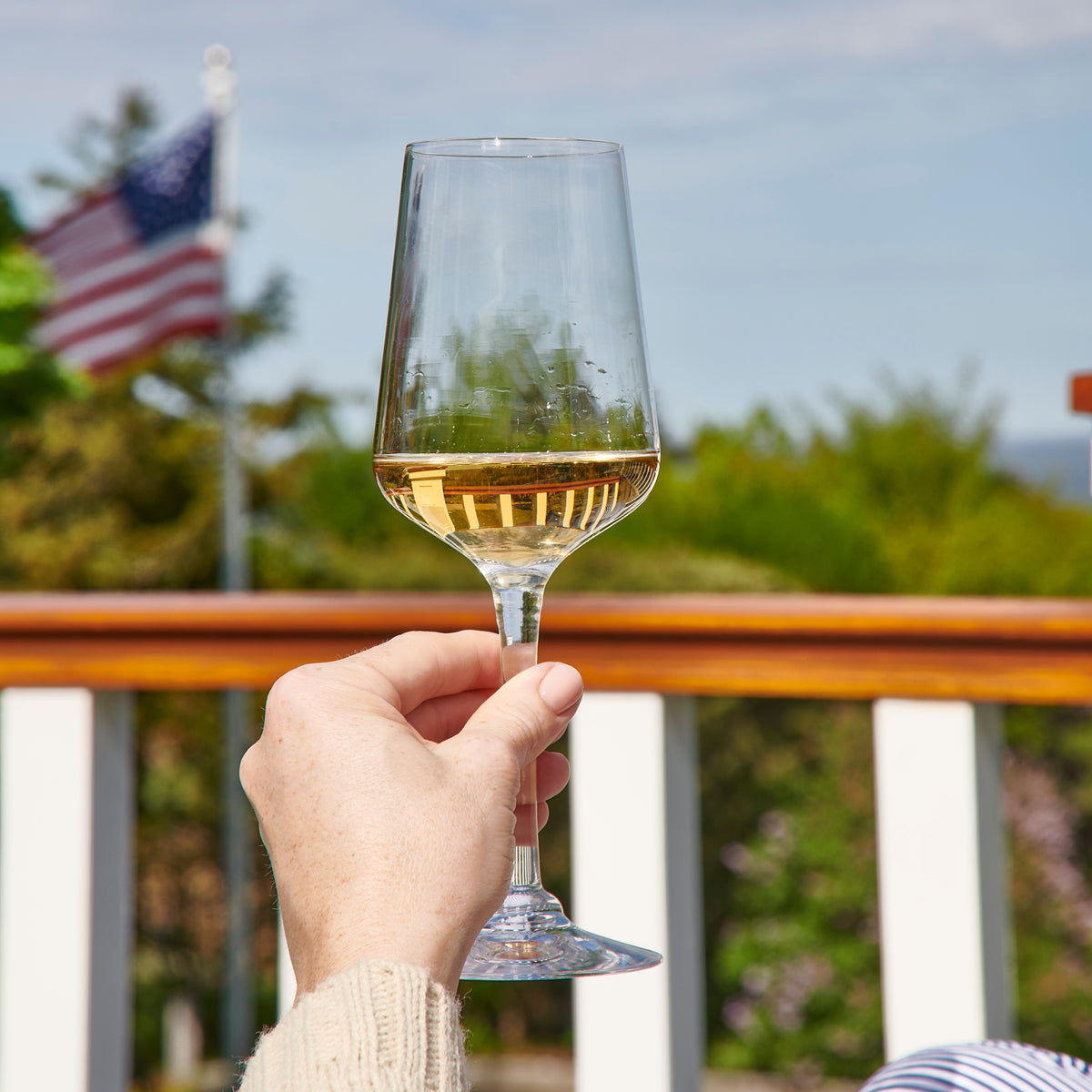 Hand holding a wine glass filled with white wine outdoors with green landscape and American flag in background.