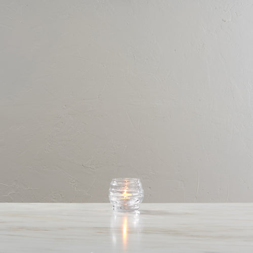 Clear glass tealight holder with lit candle inside, textured surface.