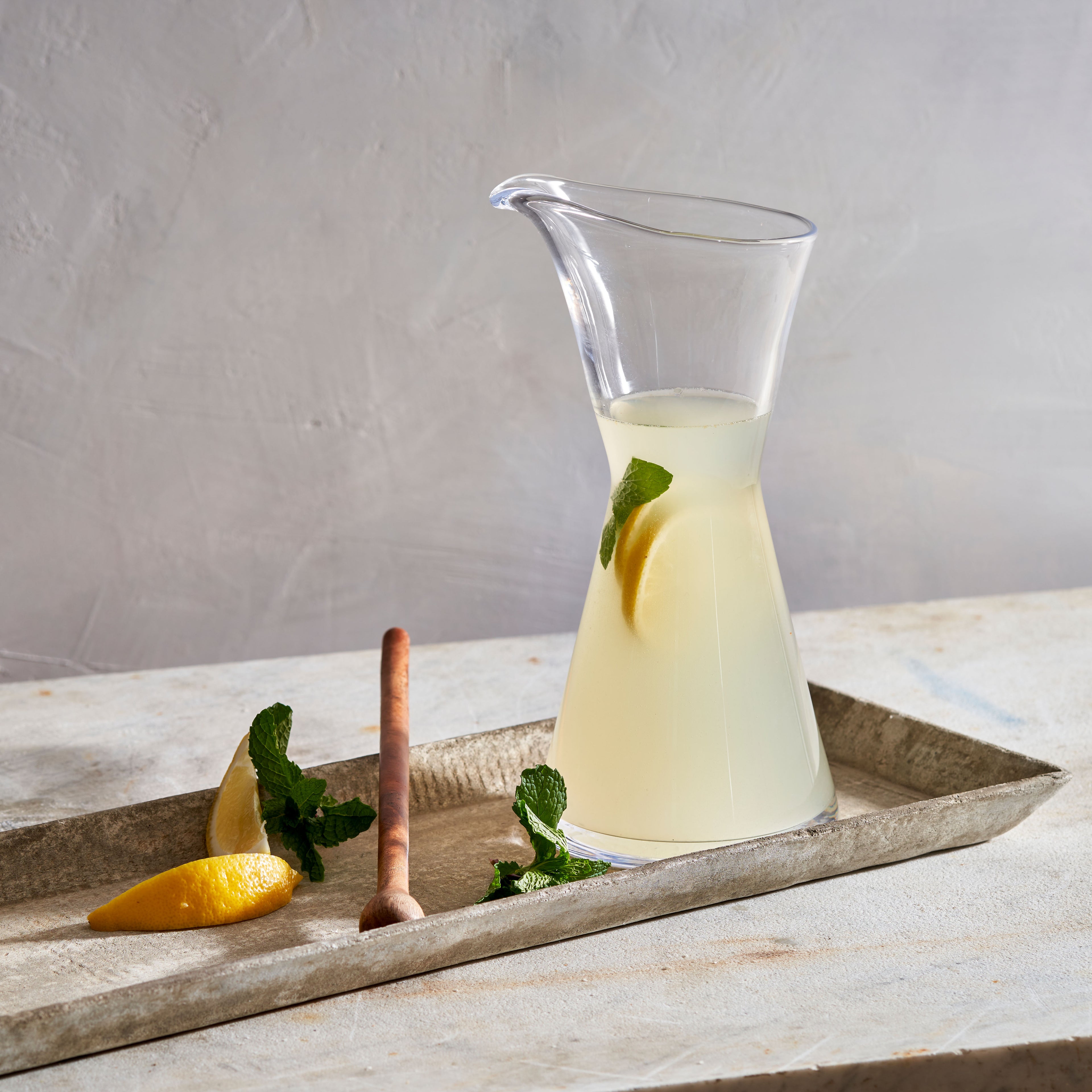Clear glass pitcher filled with pale yellow liquid, lemon slice, and mint sprig inside, displayed on concrete tray with lemon