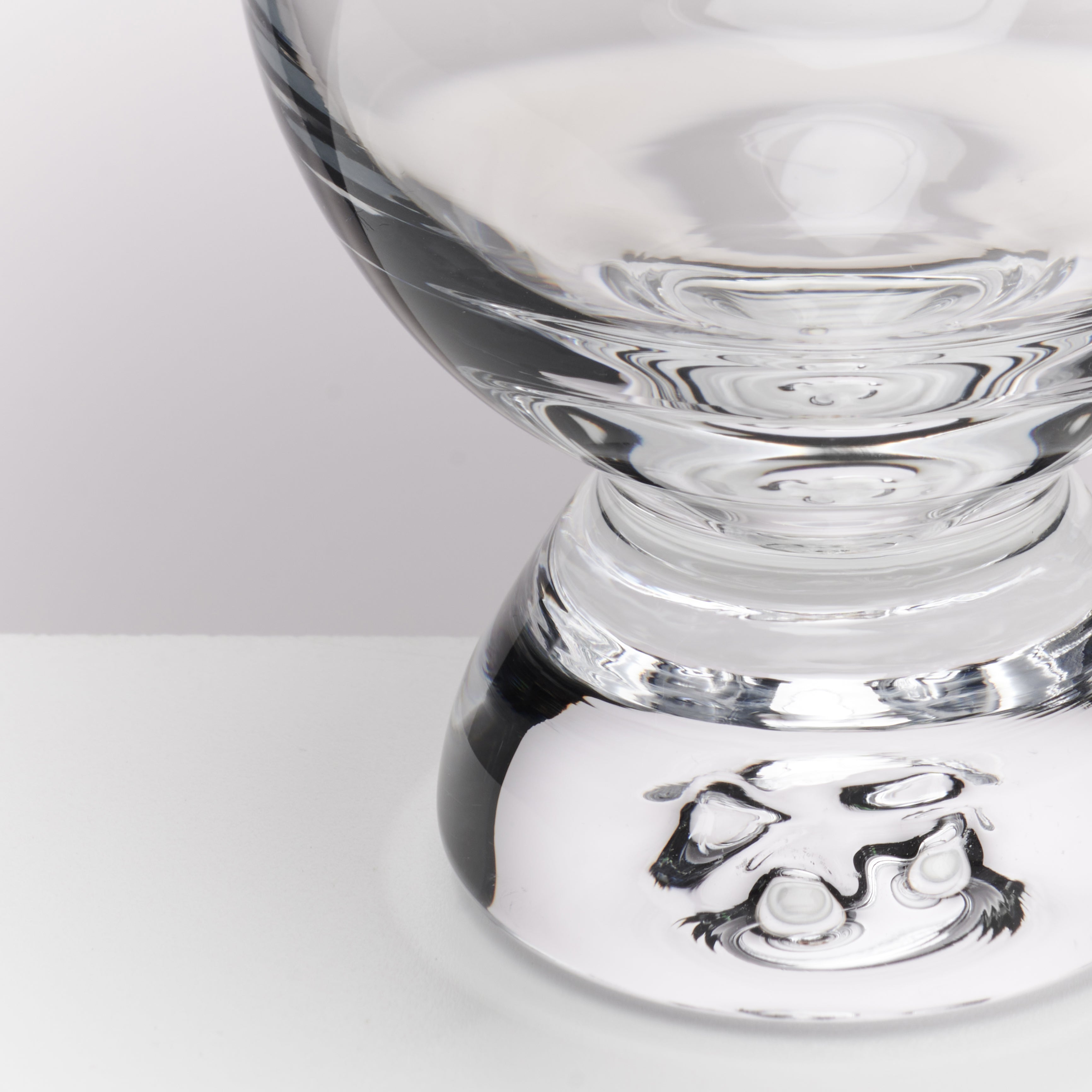 Close-up of a clear glass pedestal lamp base with a sculptural, organic-shaped bottom, photographed on a clean white surface against a light gray background.