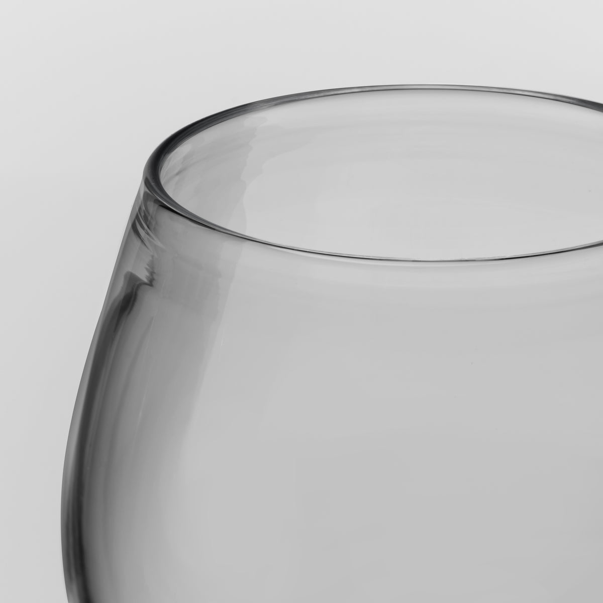Close-up of a clear glass bowl or tumbler with a thick, rounded base and a smooth, curved rim, shown against a neutral light gray background.