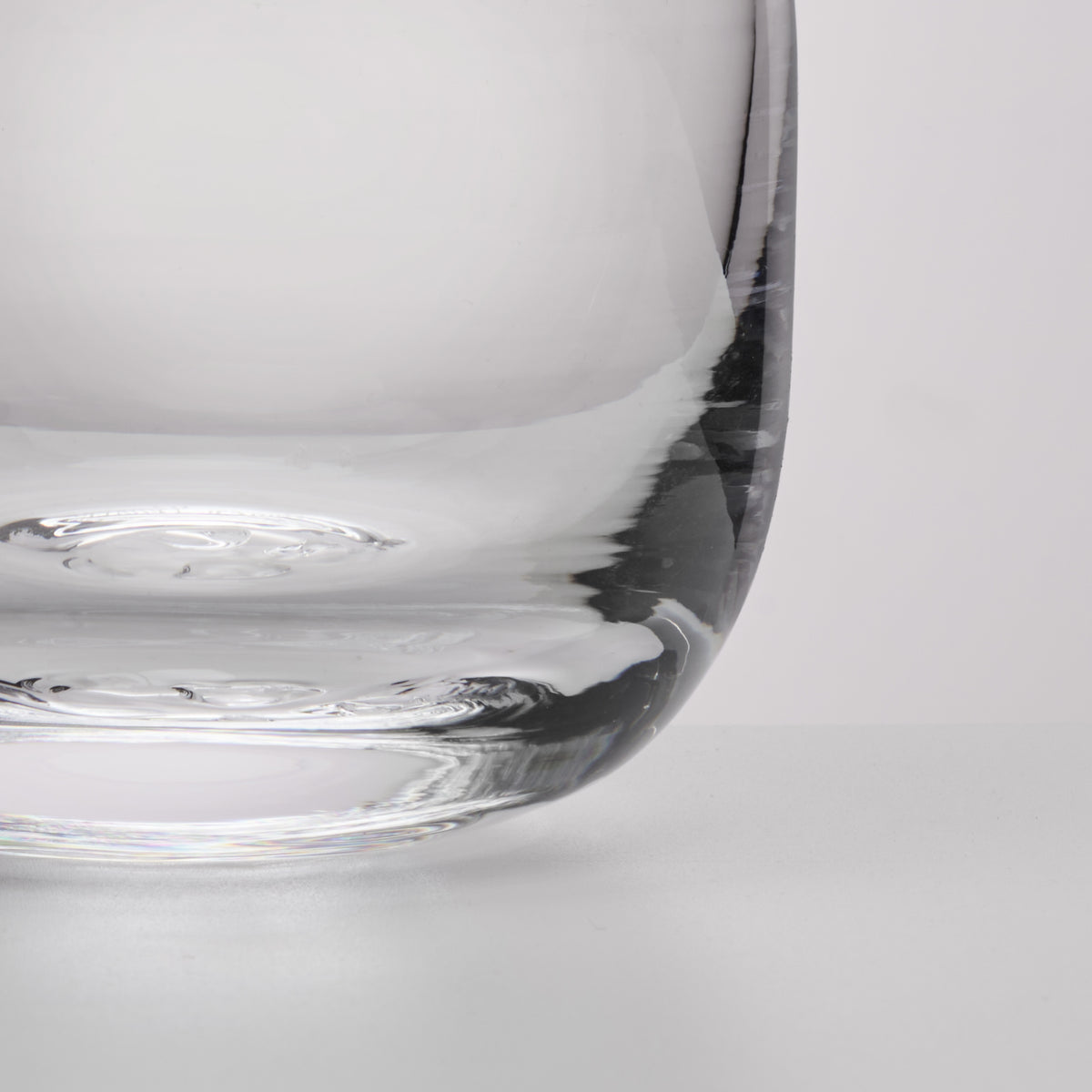 A close-up studio shot of a clear glass tumbler, showing the curved base and a portion of the transparent body against a light gray background.