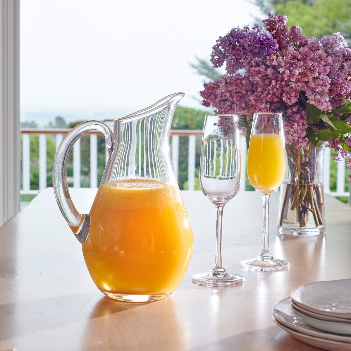 Clear glass pitcher with vertical ribbed pattern filled with orange juice, displayed on table with glasses and purple flowers