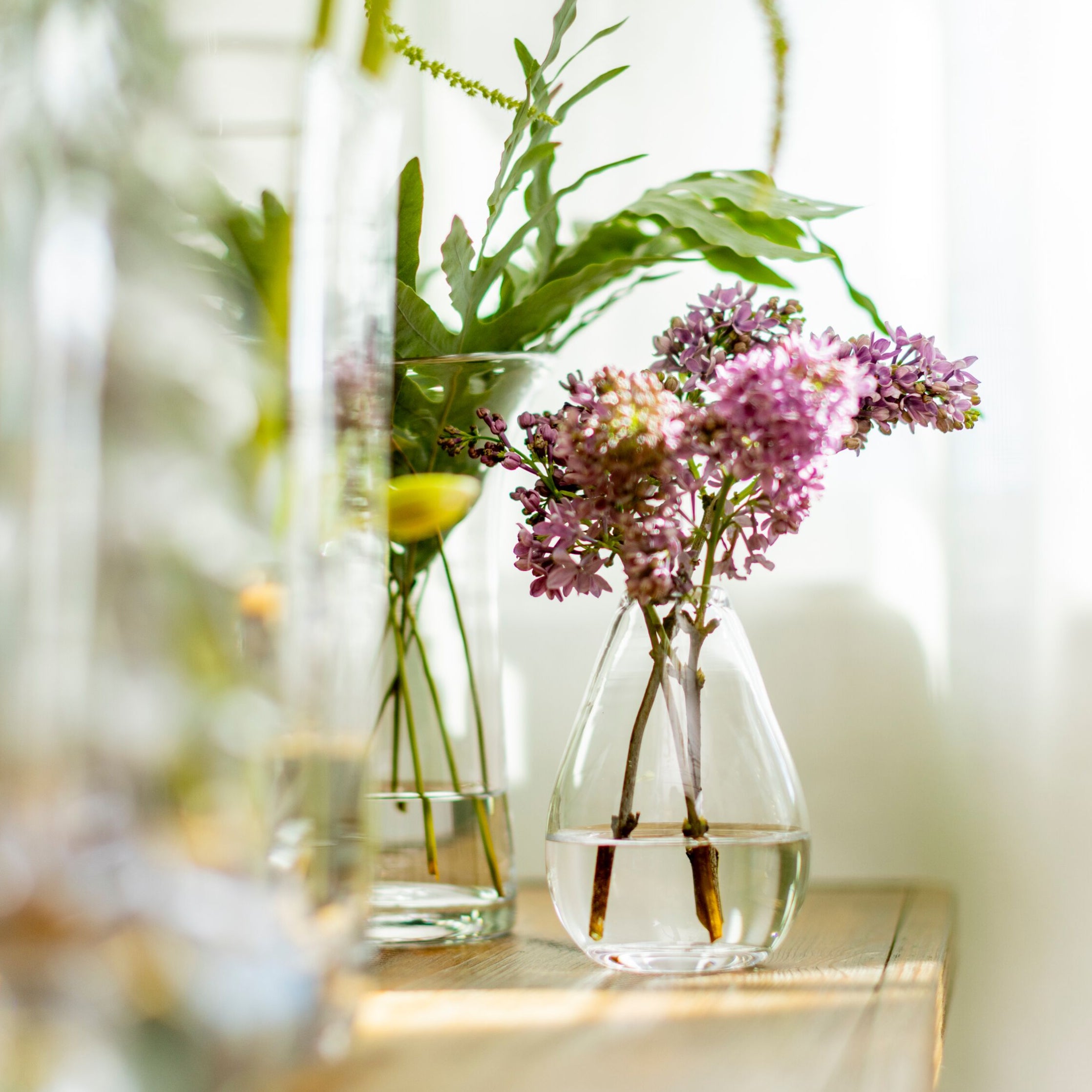 Clear glass teardrop vase holding pink and purple clustered flowers with green stems and foliage.