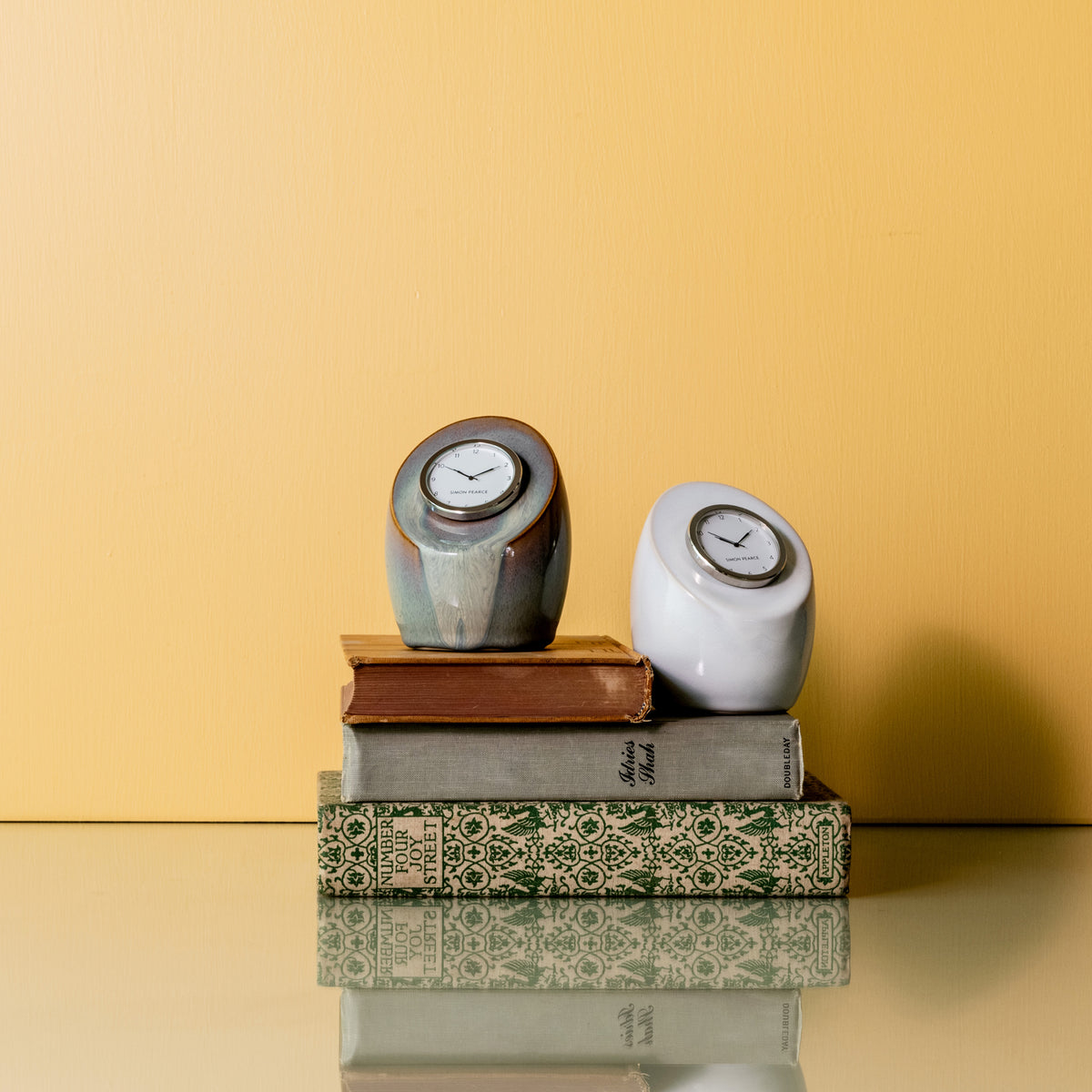 Gray and white ceramic clocks with clock faces displayed on stacked decorative books against a yellow background.