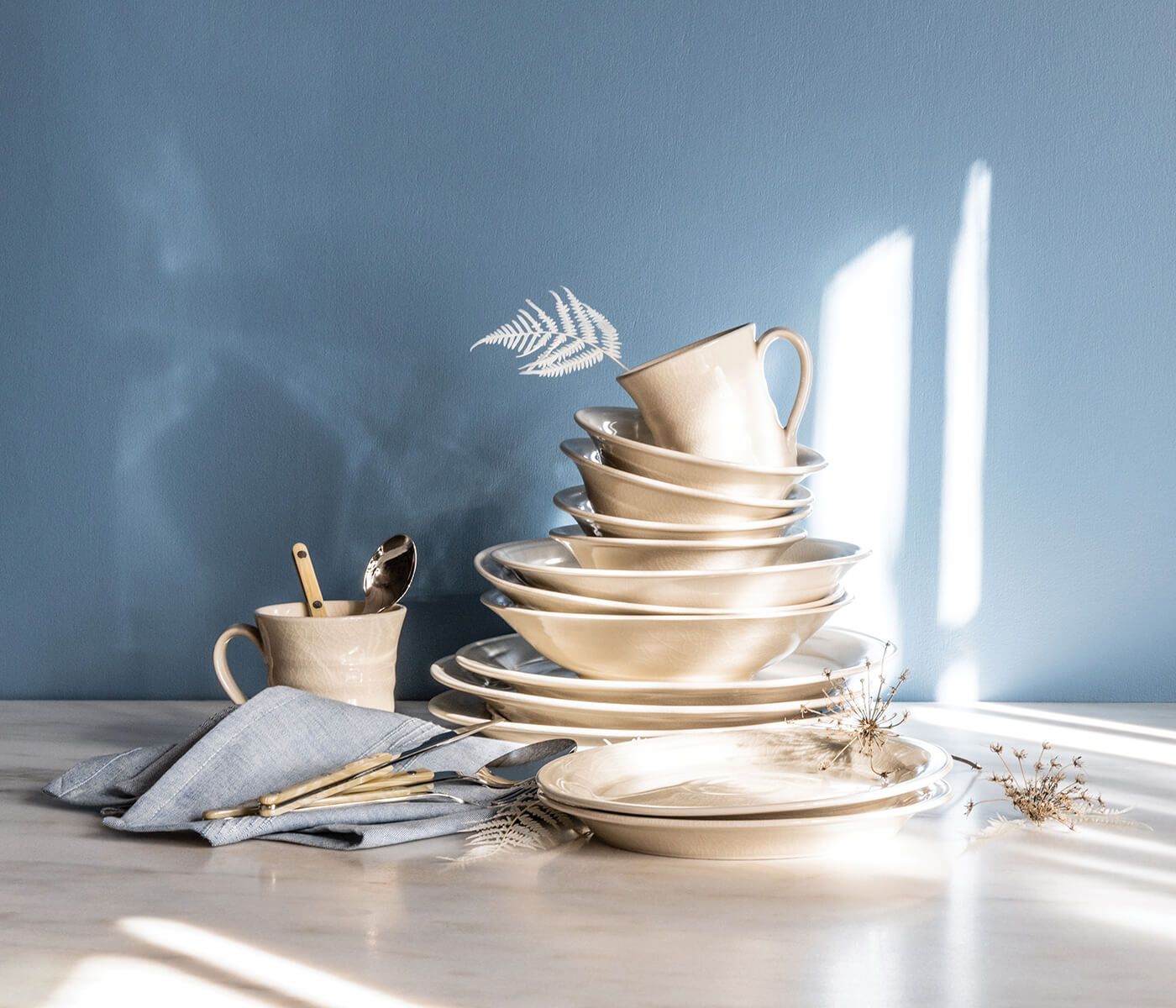 Stacked ivory dinnerware with crackle texture, including plates, bowls, and a mug with handle, arranged with utensils and nap