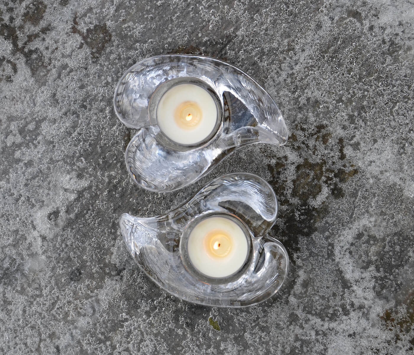 Two clear heart-shaped tealight holders with white candles and yellow flames inside, arranged vertically on concrete surface.