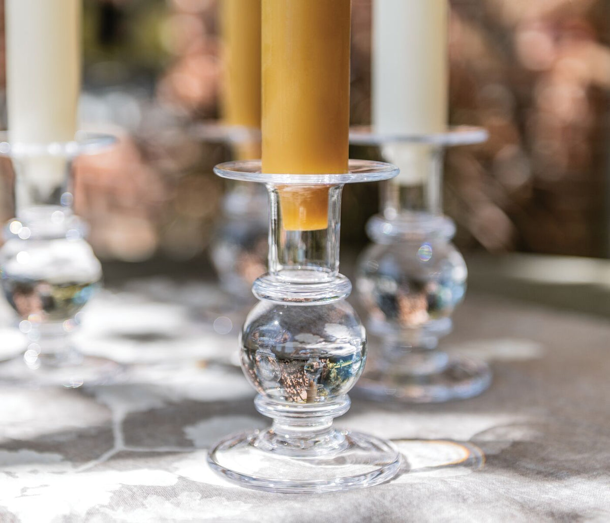 Clear glass candlestick with bulbous stem holding a tall yellow taper candle, with matching candlesticks blurred in backgroun