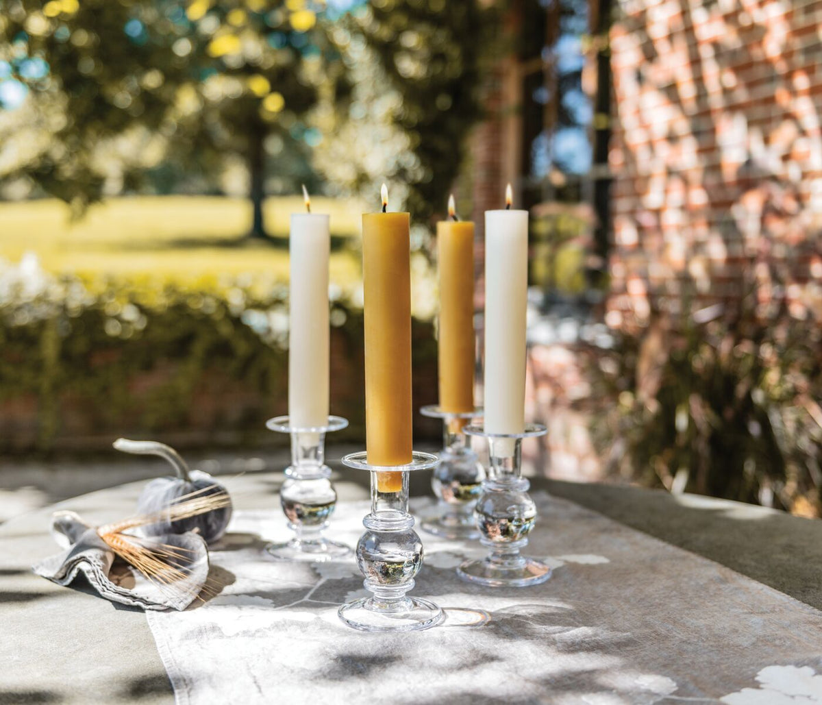 Four glass candlesticks holding white and gold tapered candles on an outdoor table.