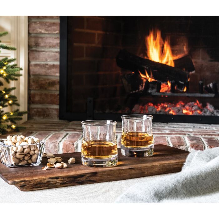 Two whiskey glasses with amber liquid and a bowl of nuts on a wooden tray by a fireplace.