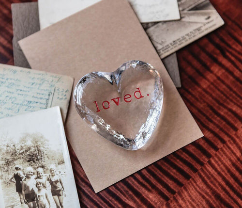Clear glass heart-shaped object with "loved" text etched in red, displayed on tan paper.