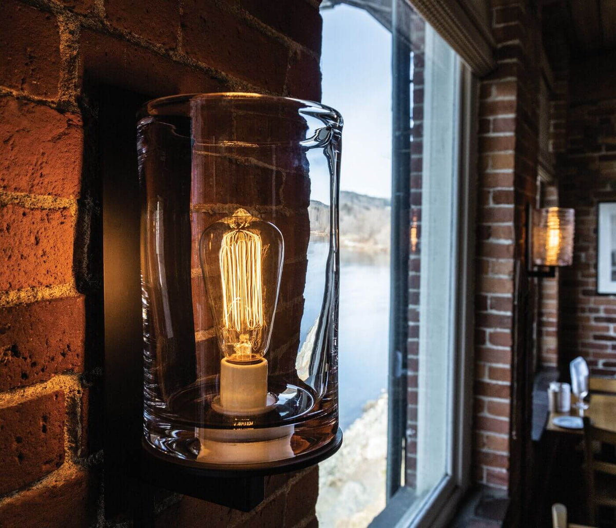 Clear glass cylindrical sconce with Edison-style filament bulb mounted on brick wall.