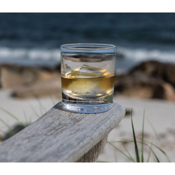 Glass of whiskey with ice on a wooden surface by a beach.