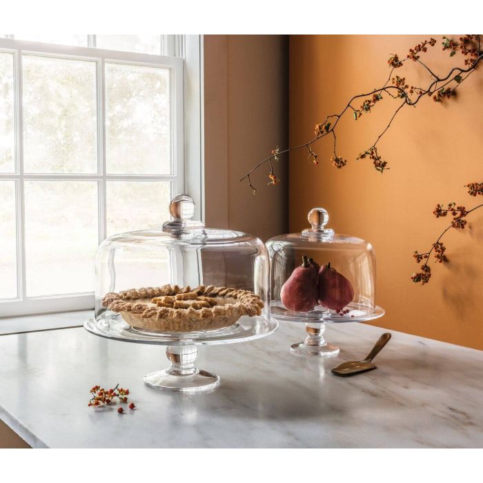 Two clear glass cake plates with domed lids on pedestal bases, one holding a pie and one with fruit inside.