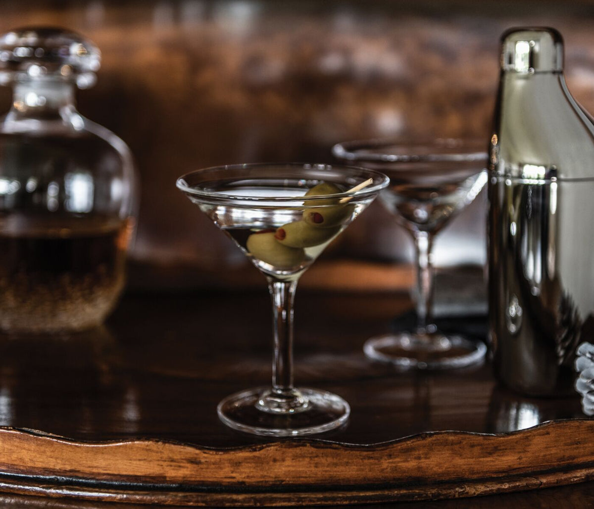 Two martini glasses with olives on a pick, cocktail shaker, and bottle on a wooden surface.