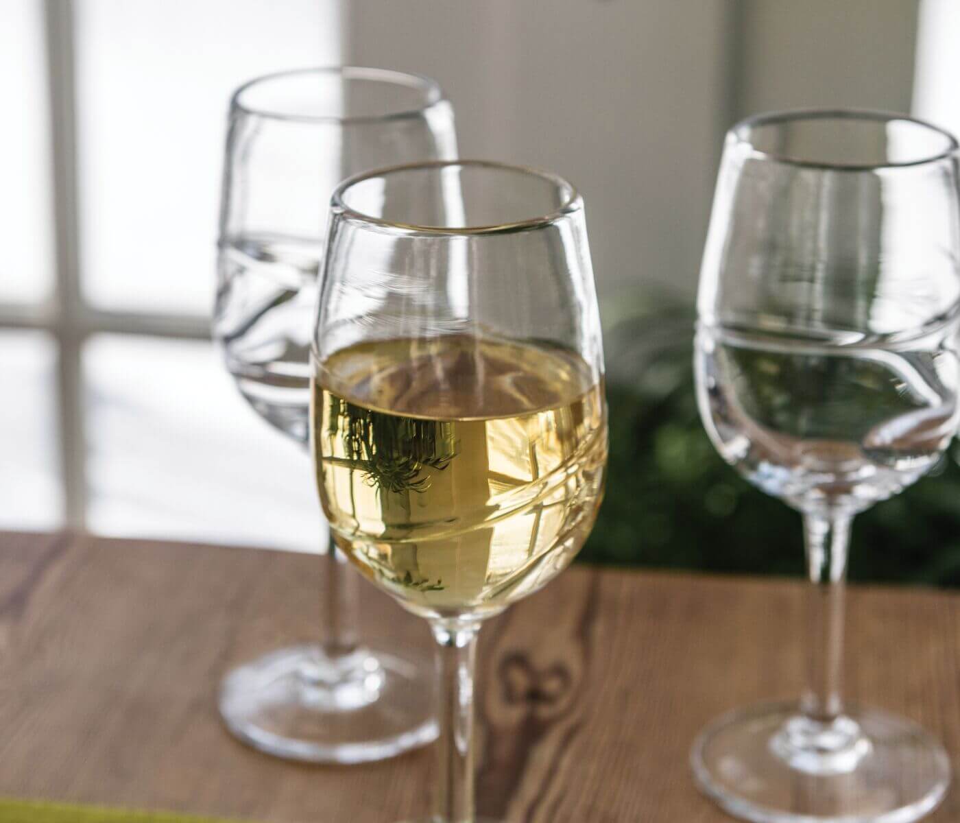 Three wine glasses on wooden surface, center glass filled with pale yellow wine, two empty glasses beside it.