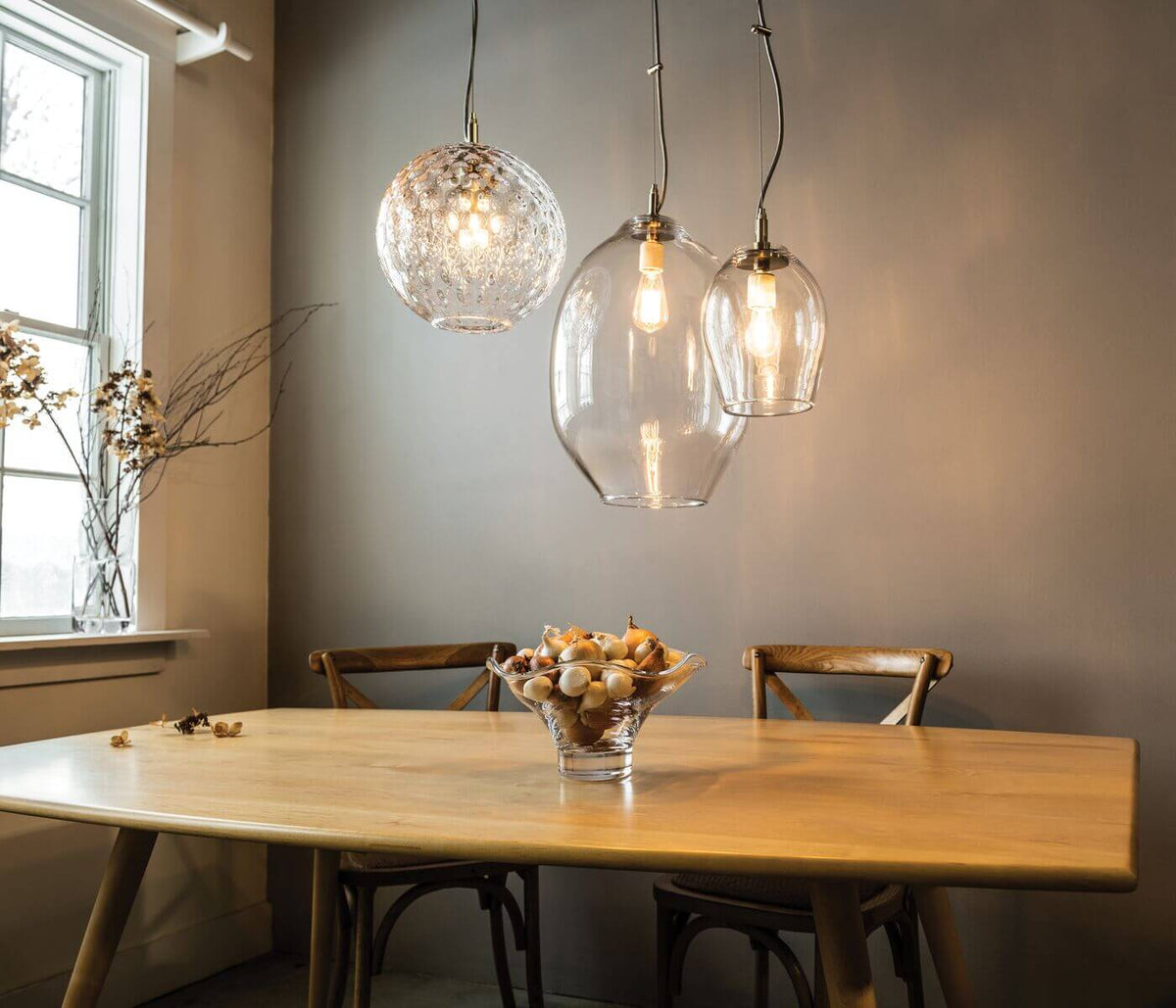 Three glass pendant lights with brass fittings hang above a wooden dining table, featuring textured and clear globe shapes.