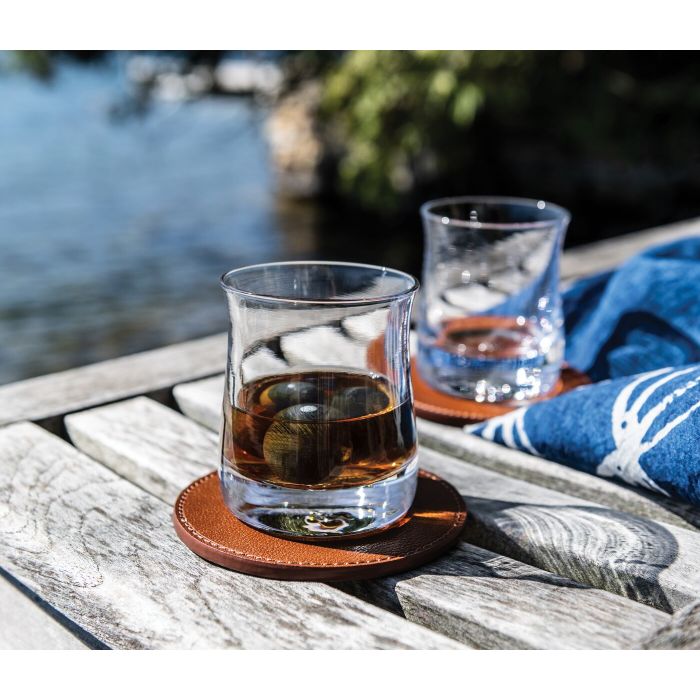 Two whiskey glasses with ice on brown leather coasters beside a second empty glass on weathered wood dock.