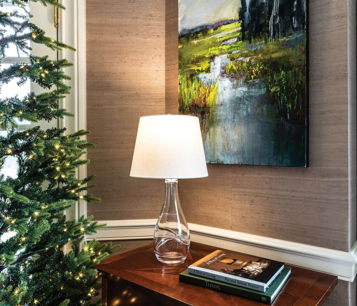Clear glass bottle-form lamp with white fabric shade on wooden table next to books.