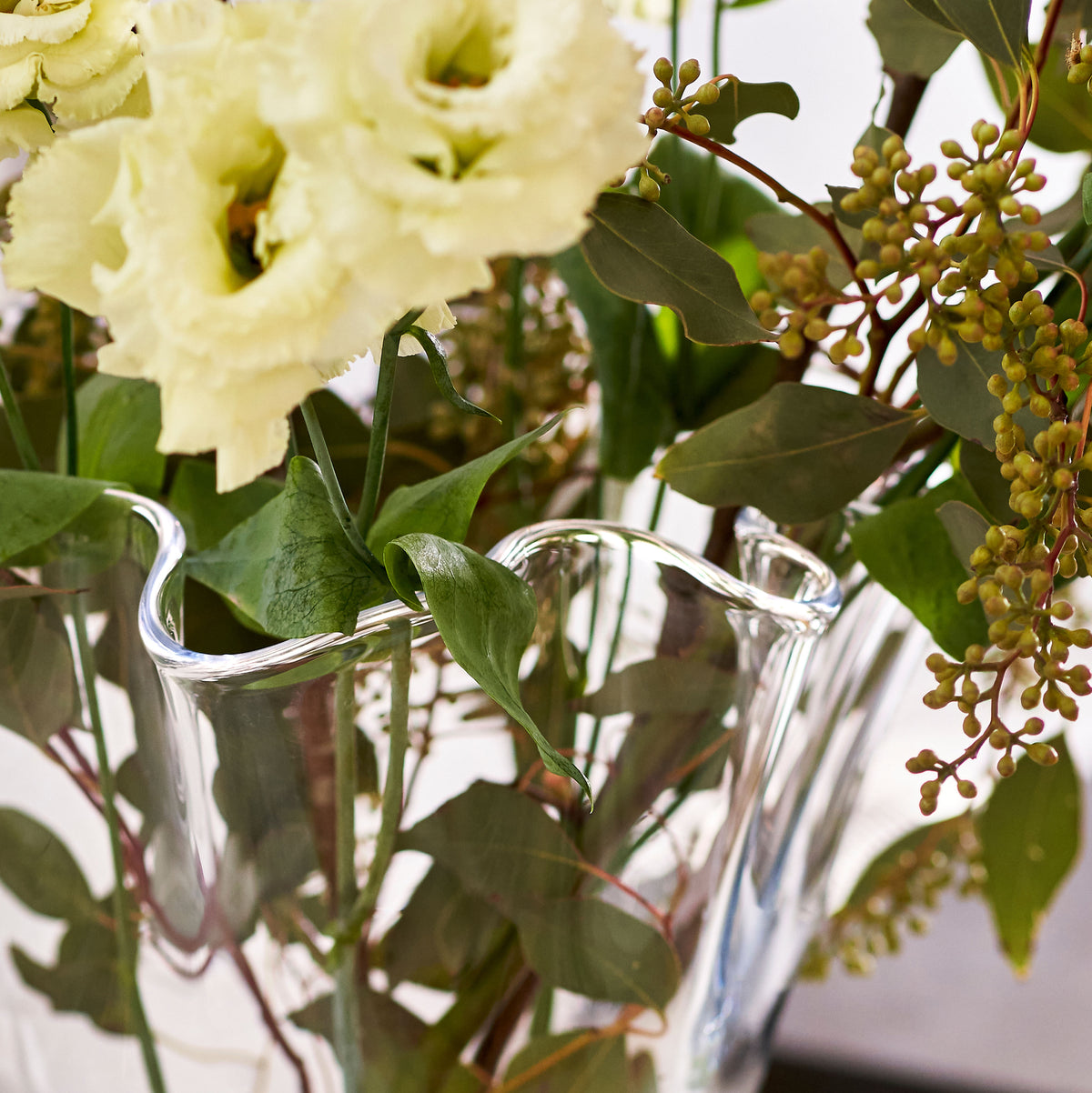 Clear glass vase with white anemone flowers, green foliage, and yellow-brown berry clusters arranged inside.