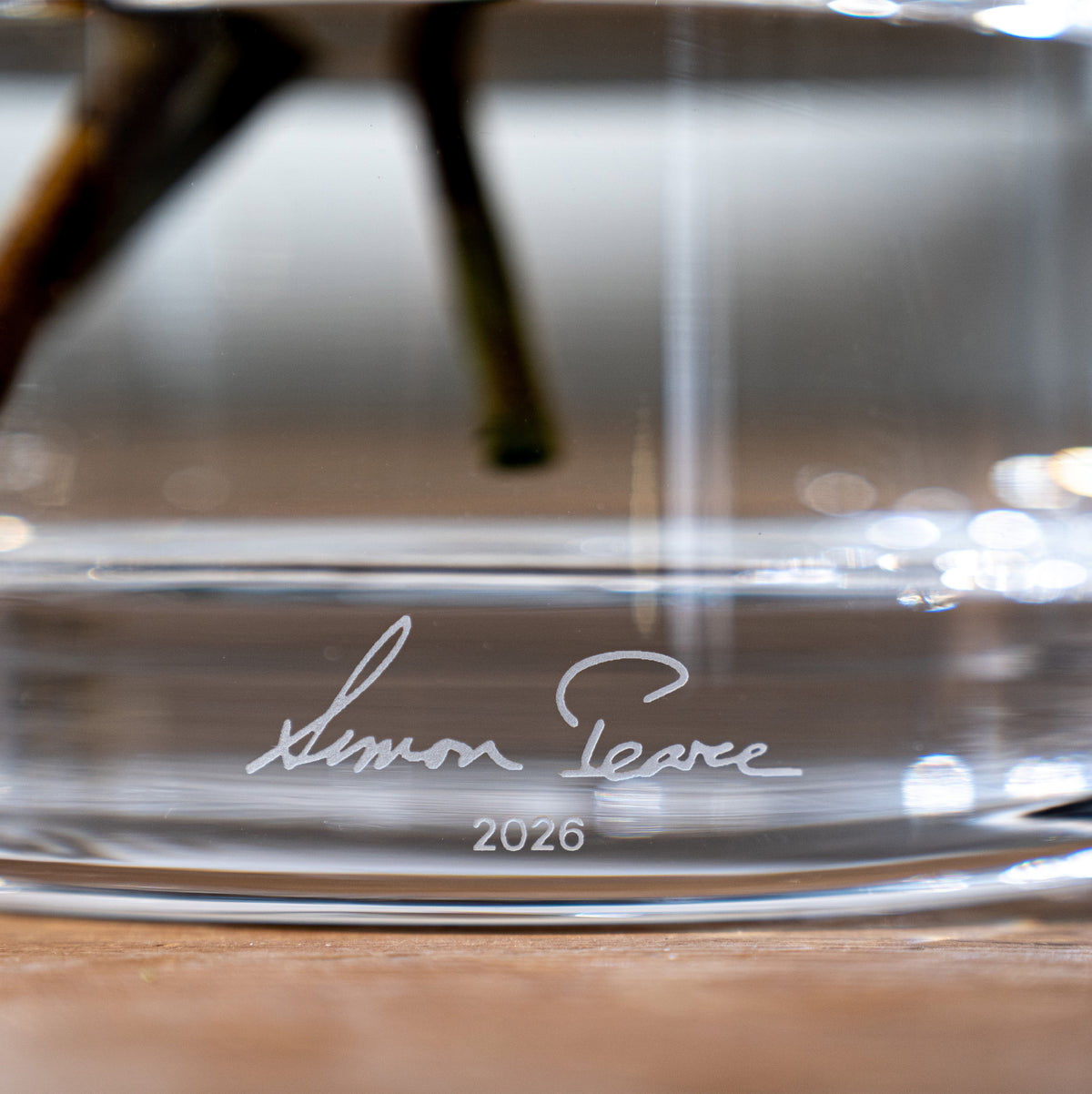 Clear glass vase with engraved signature and "2026" on the base, shown from a close-up angle.