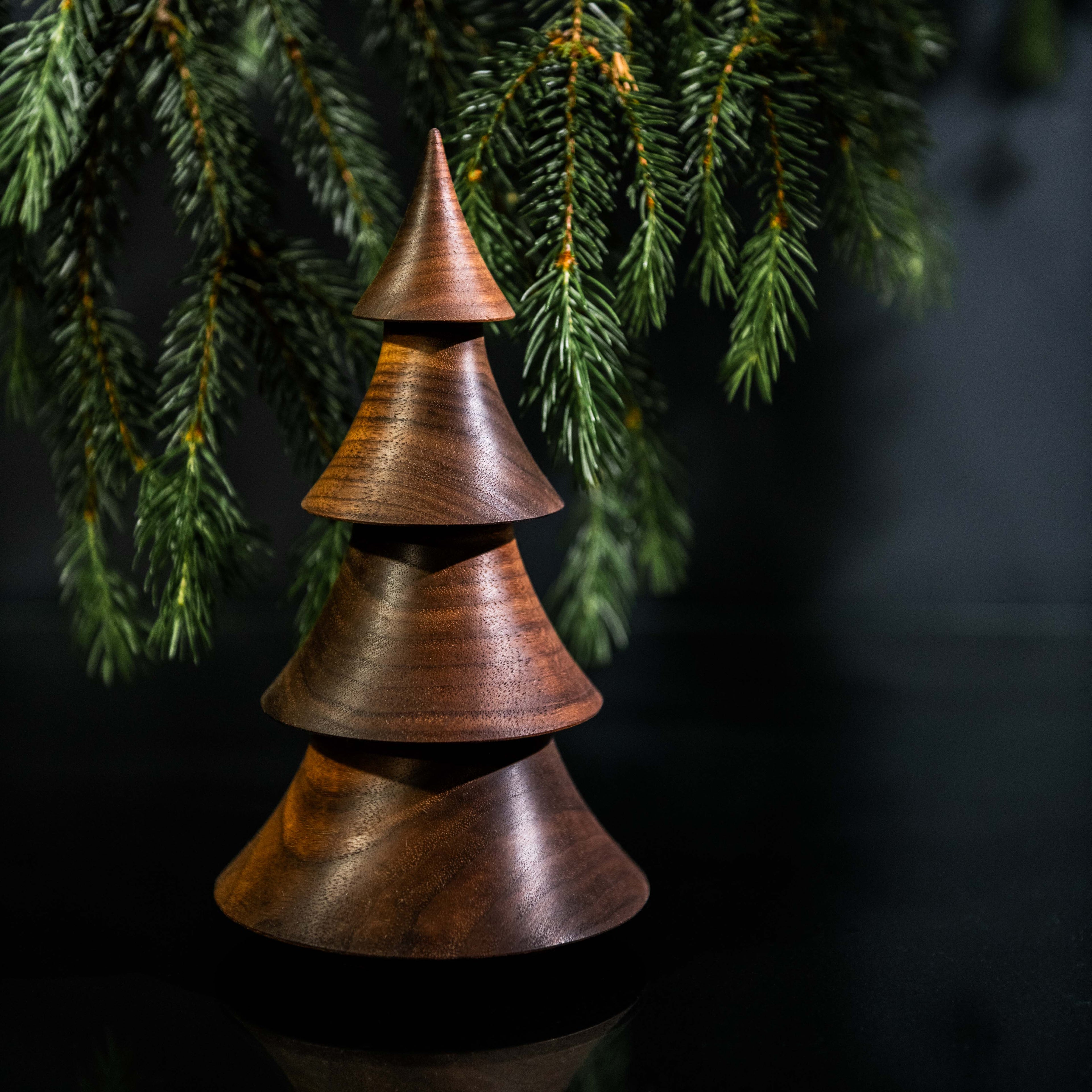 Stacked walnut wood tree-shaped ornament with pointed top beneath evergreen branches.