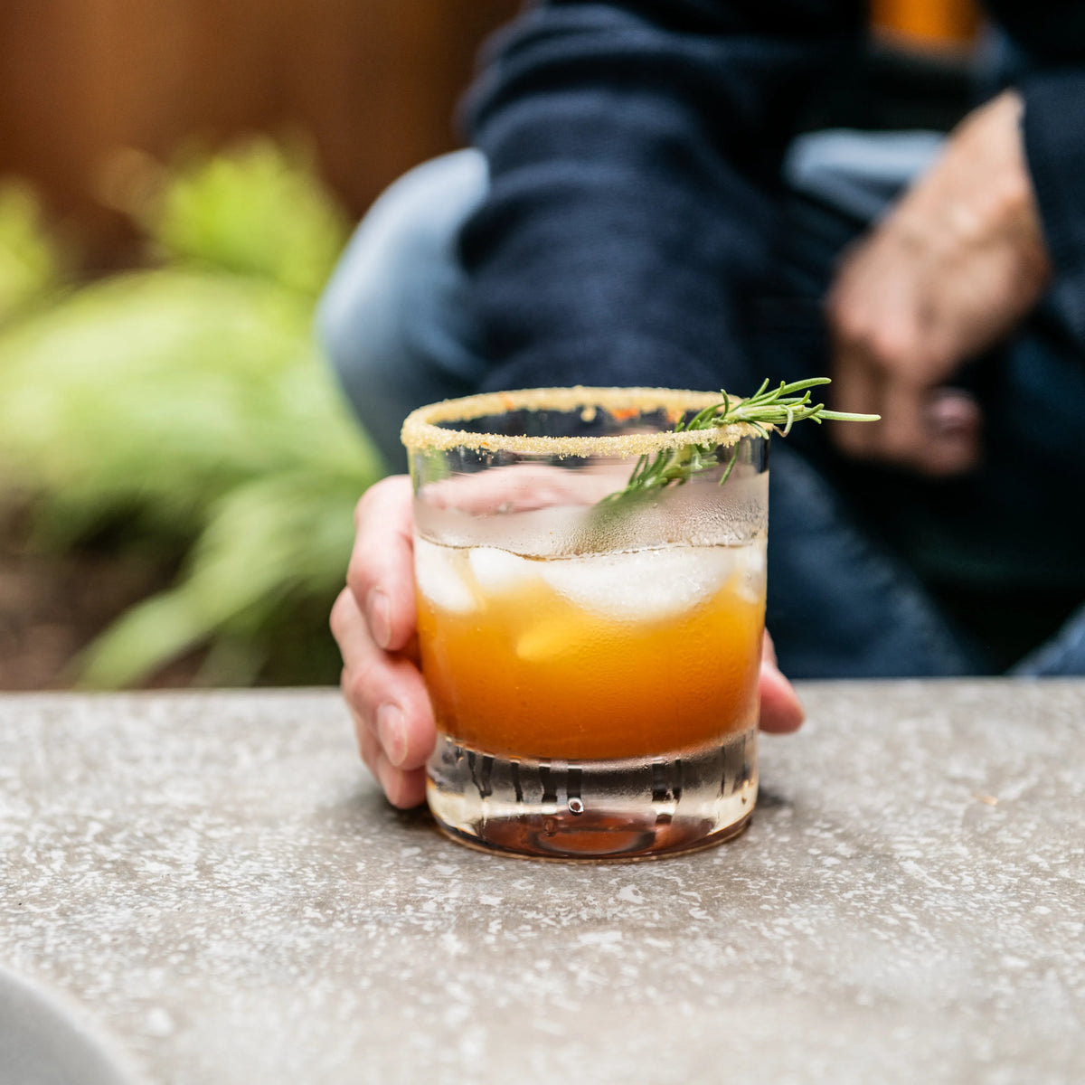 Double old-fashioned glass filled with amber liquid and ice, garnished with rosemary sprig, held in hand.