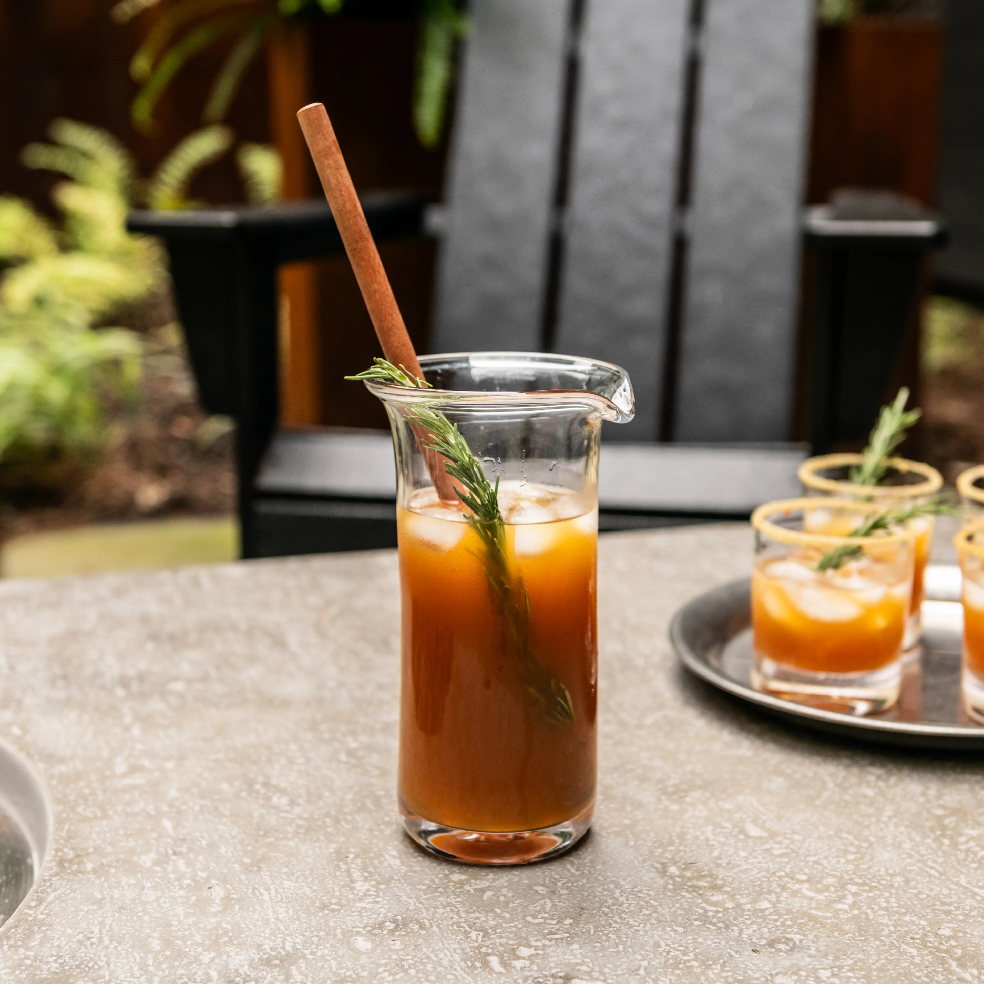 Clear glass pitcher filled with brown liquid, wooden stirrer, and fresh herb garnish on outdoor table.