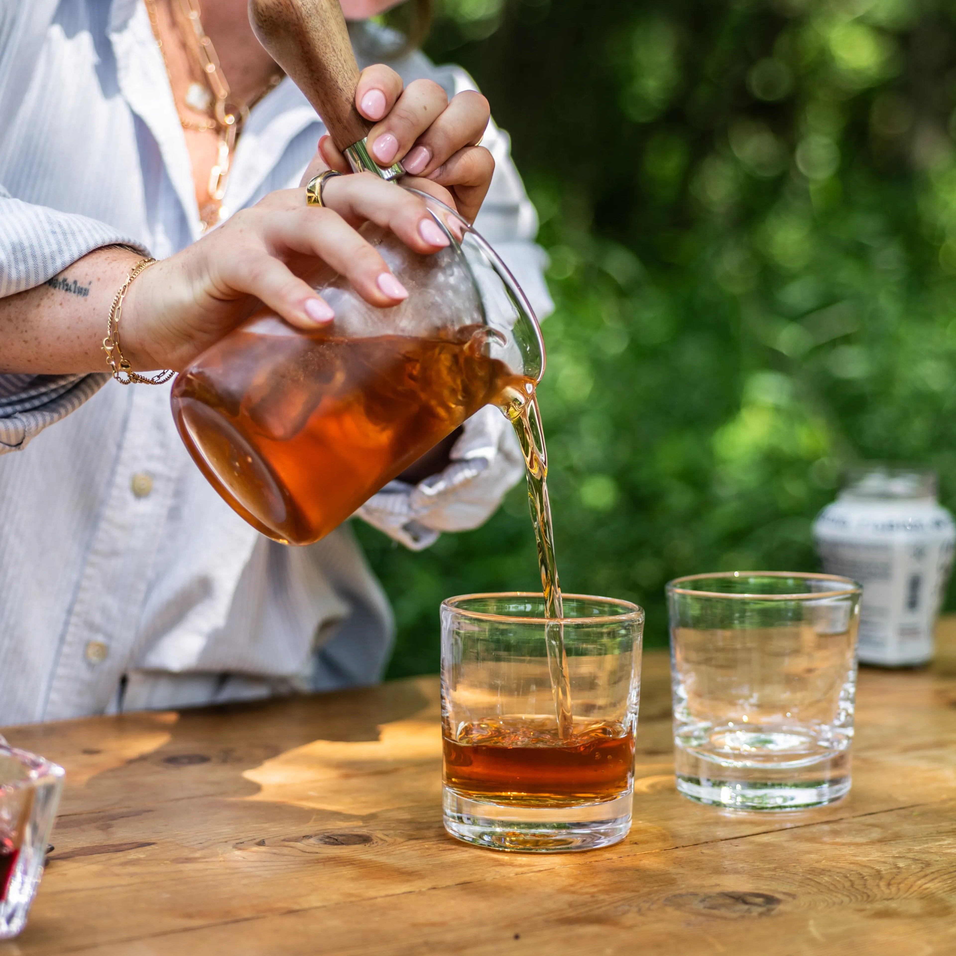 Glass pitcher with gold-tone handle pouring amber liquid into rocks glasses outdoors.