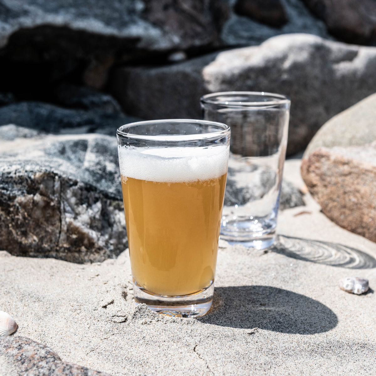 Two clear pint glasses, one filled with amber beer and one empty, set on sand with rocks in background.