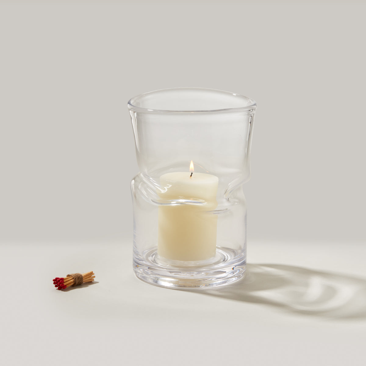 Large clear glass hurricane with cream-colored pillar candle inside and bundled matches beside it.