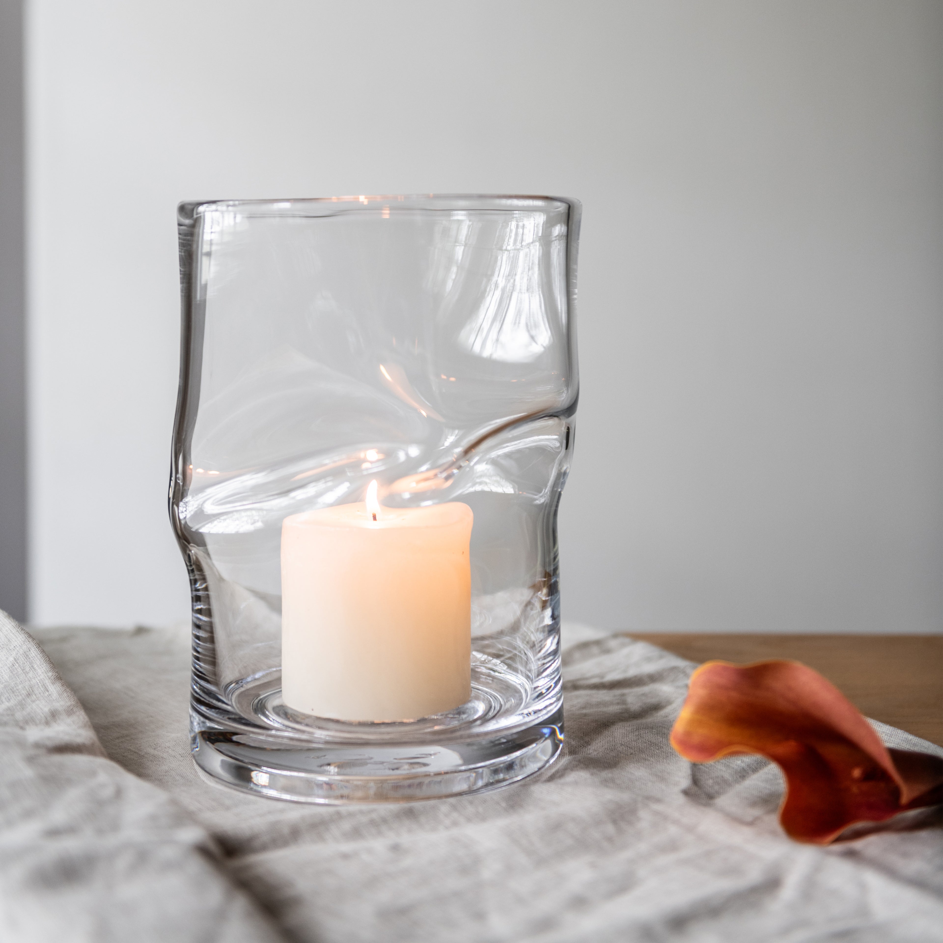 Clear glass hurricane with twisted design holding a lit cream-colored pillar candle on linen fabric.