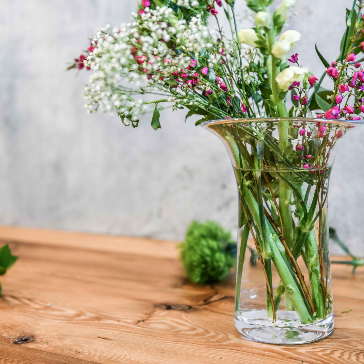 Clear glass cylinder vase with flared rim holding white baby's breath and pink flowers on wooden surface.