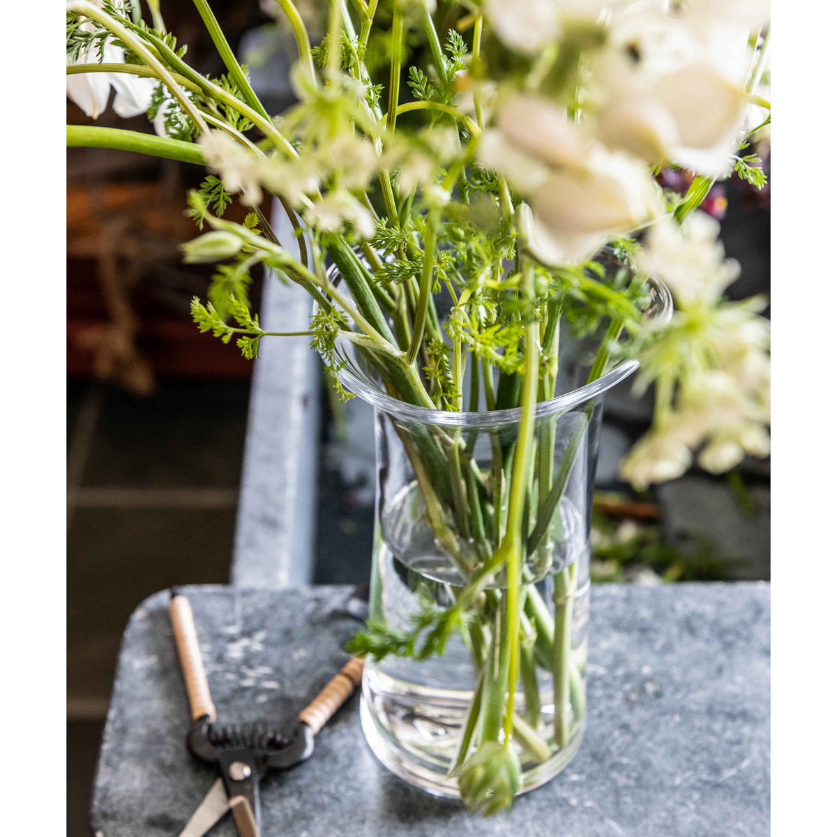 Clear glass cylindrical vase holding fresh green herbs and white flowers with wooden-handled brushes beside it.