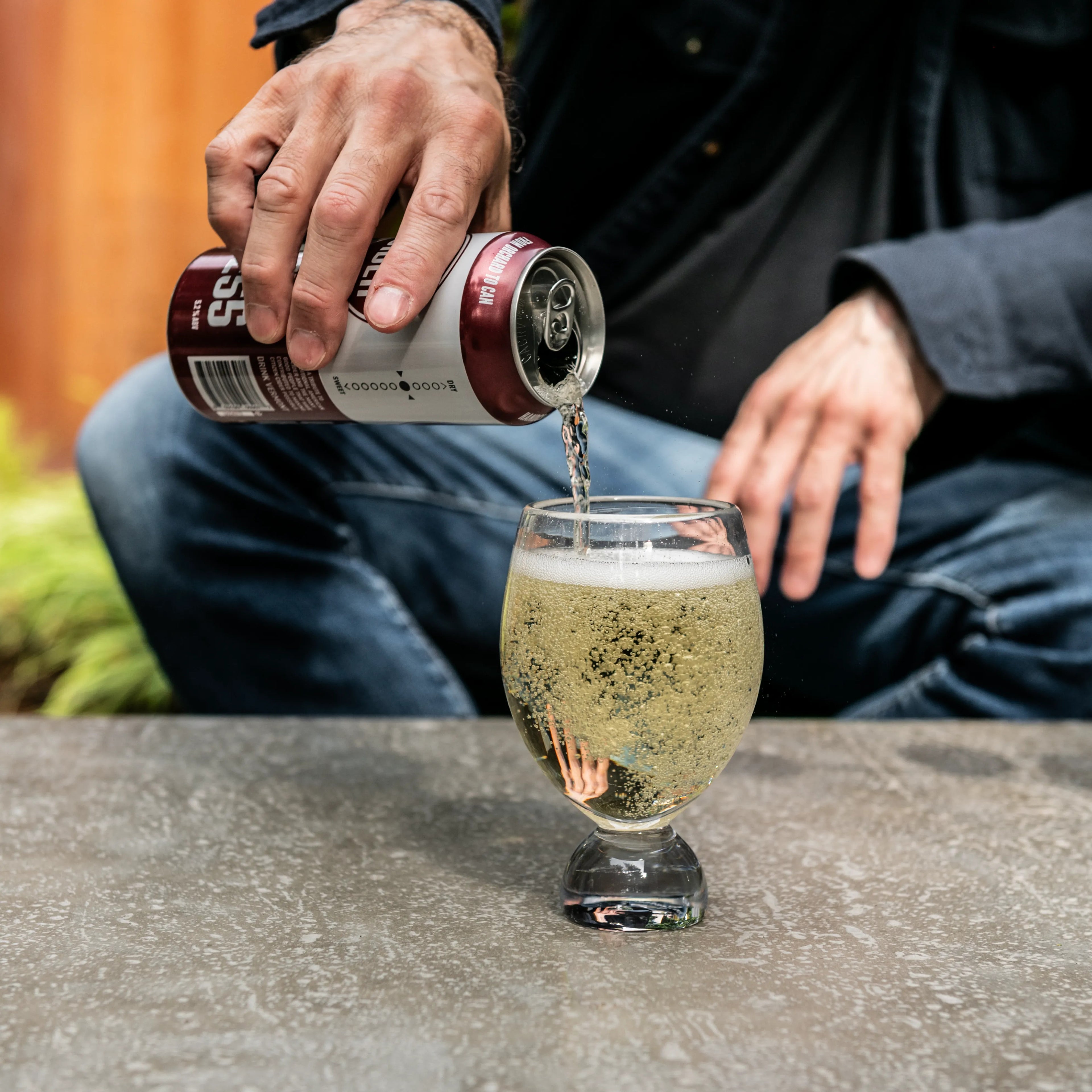 Person pouring sparkling beverage from burgundy tumbler into clear glass cup outdoors.