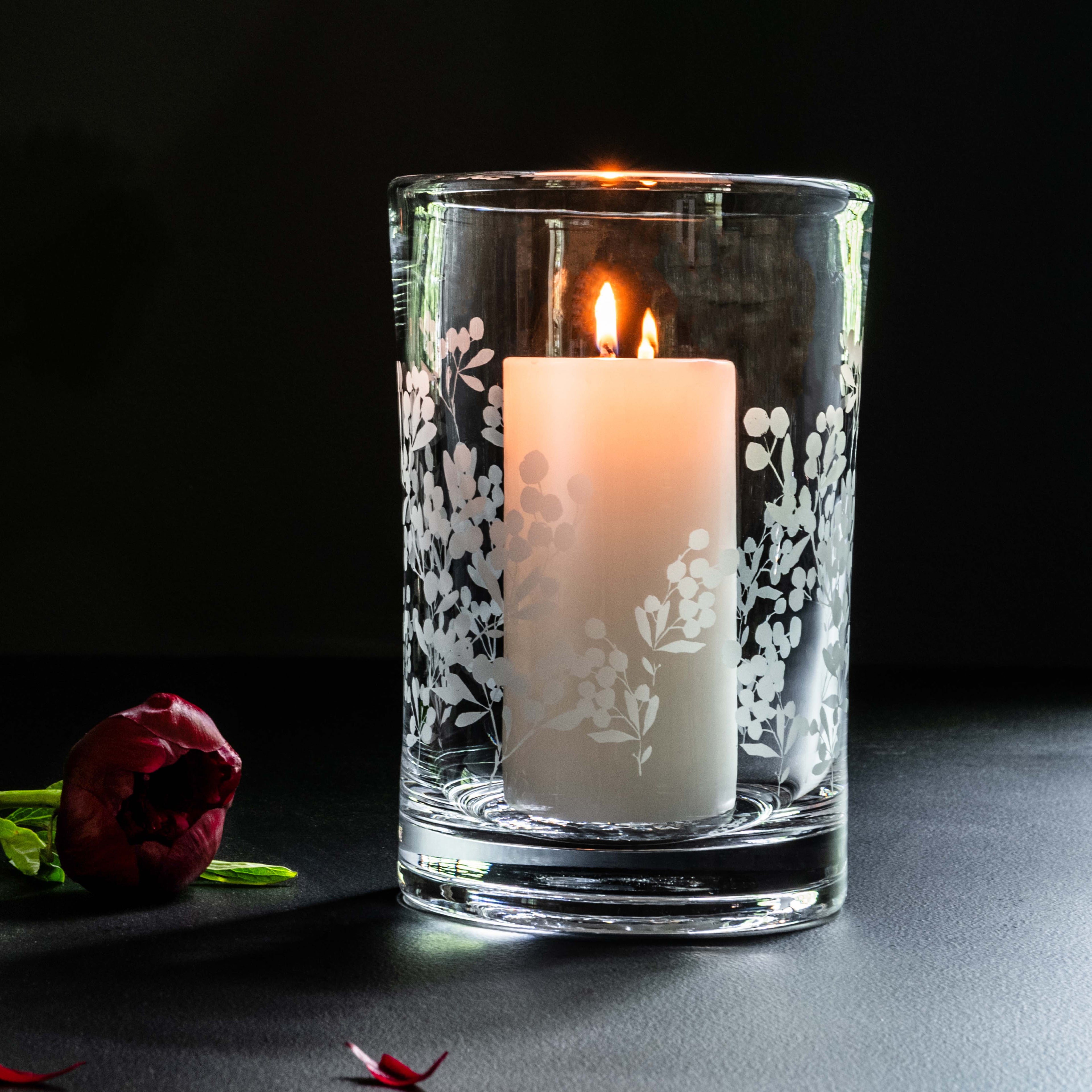 Clear glass hurricane vase with engraved berry and leaf pattern, containing a lit cream pillar candle with two flames.