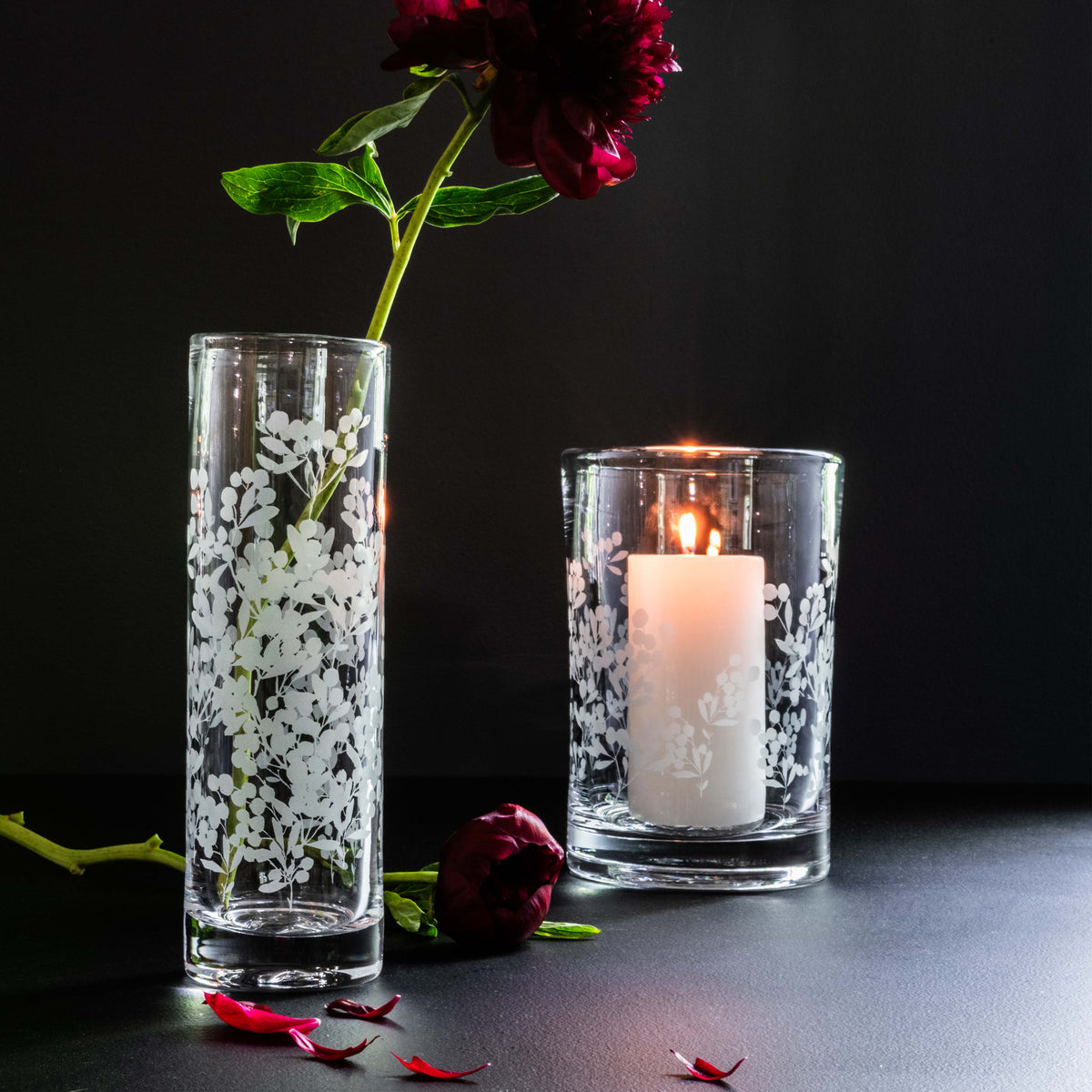 Tall and short clear glass hurricane vases with white berry and leaf engravings, one holding a red flower and the other a lit