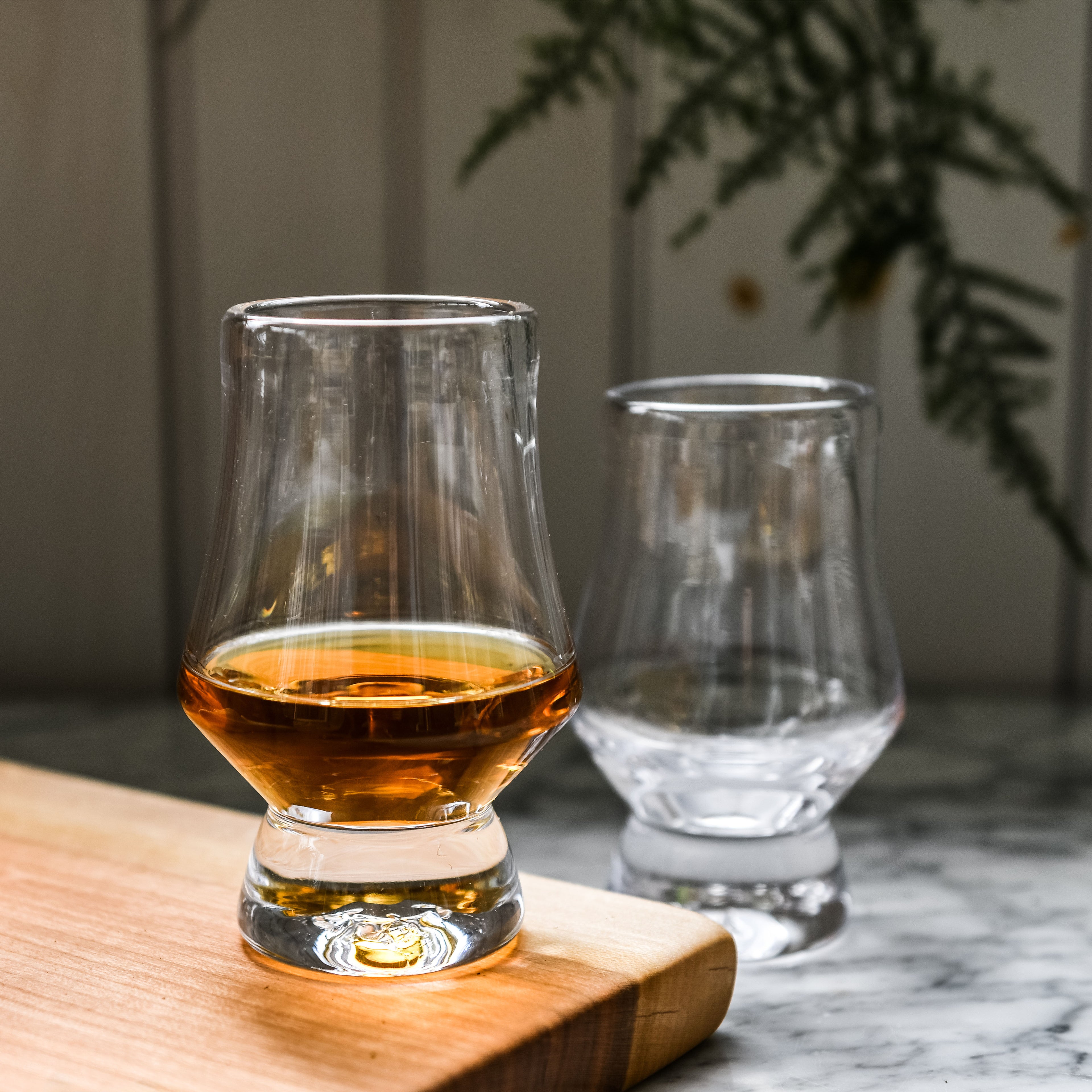 Two tapered whiskey glasses, one filled with amber bourbon and one empty, displayed on a wooden surface.