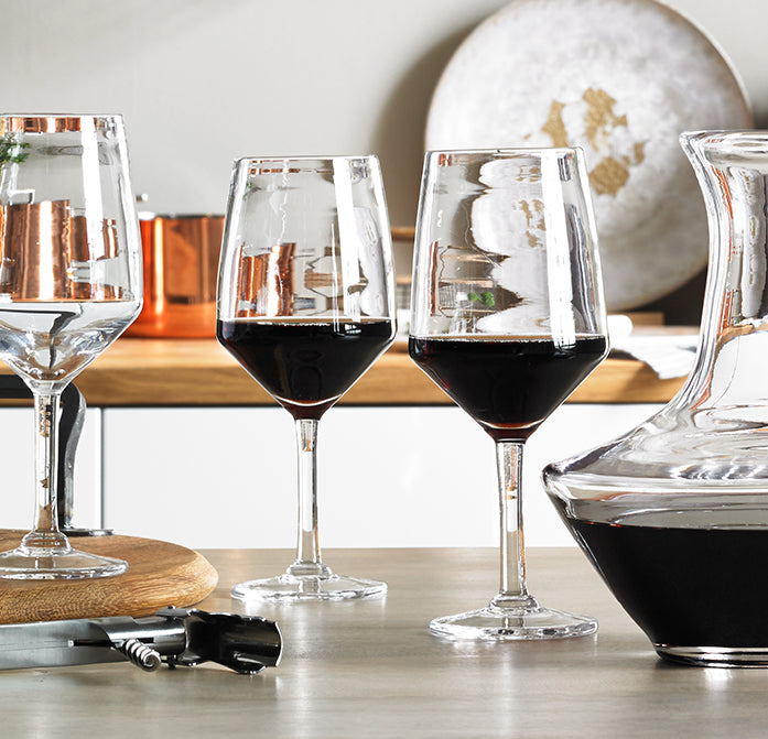 Two glasses filled with dark red wine on a wooden surface with wine bottles and accessories.