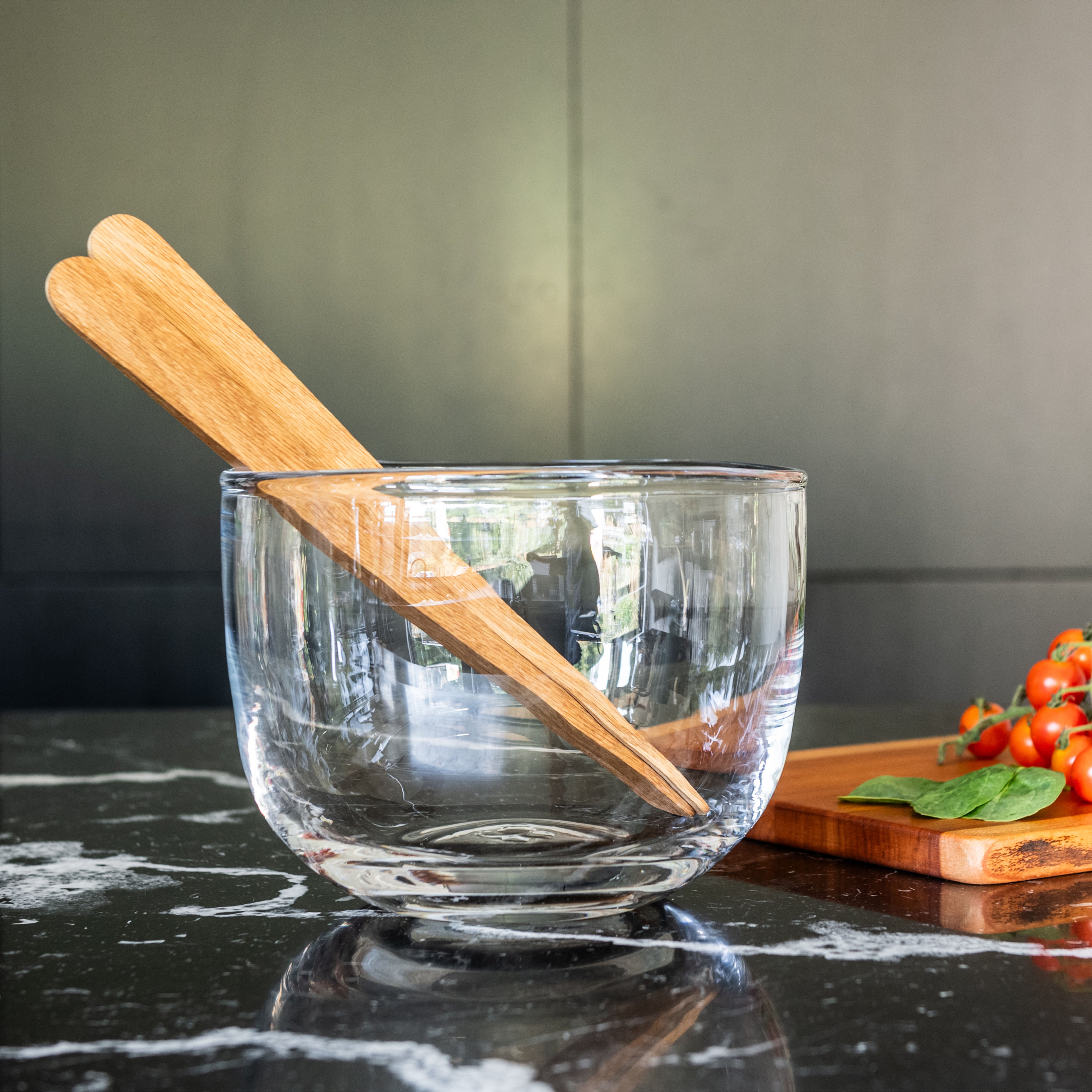 Clear glass bowl with wooden pestle inside, placed on dark countertop next to cutting board with tomatoes and basil.