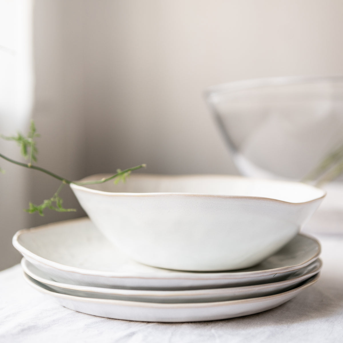 Stacked white dinnerware plates and bowls with curved edges, styled with green plant sprigs.