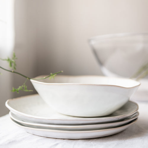 Stacked white dinnerware plates and bowls with curved edges, styled with green plant sprigs.