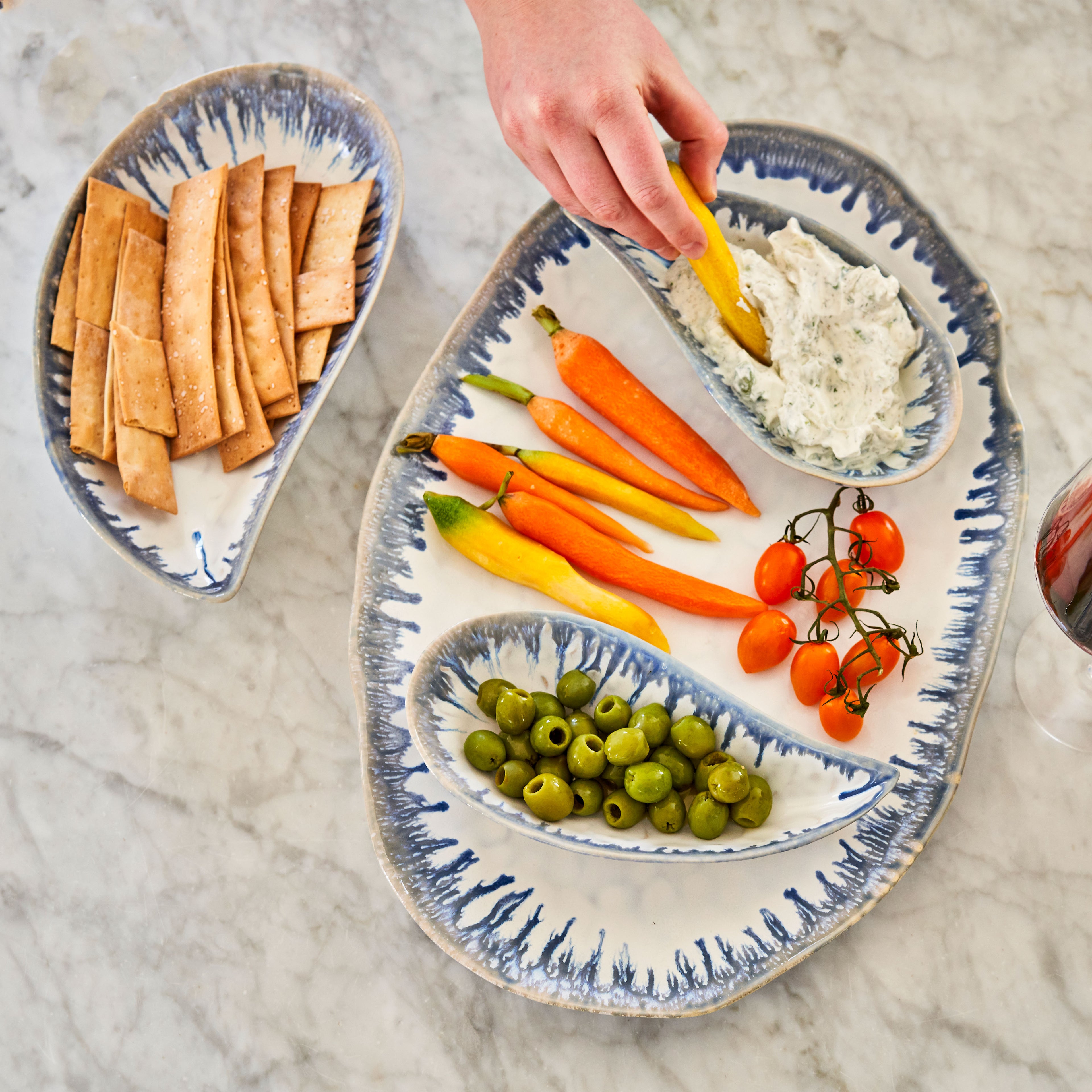 Blue and white striped mussel shell-shaped serving bowls holding crackers, vegetables, olives, and dip on marble surface.