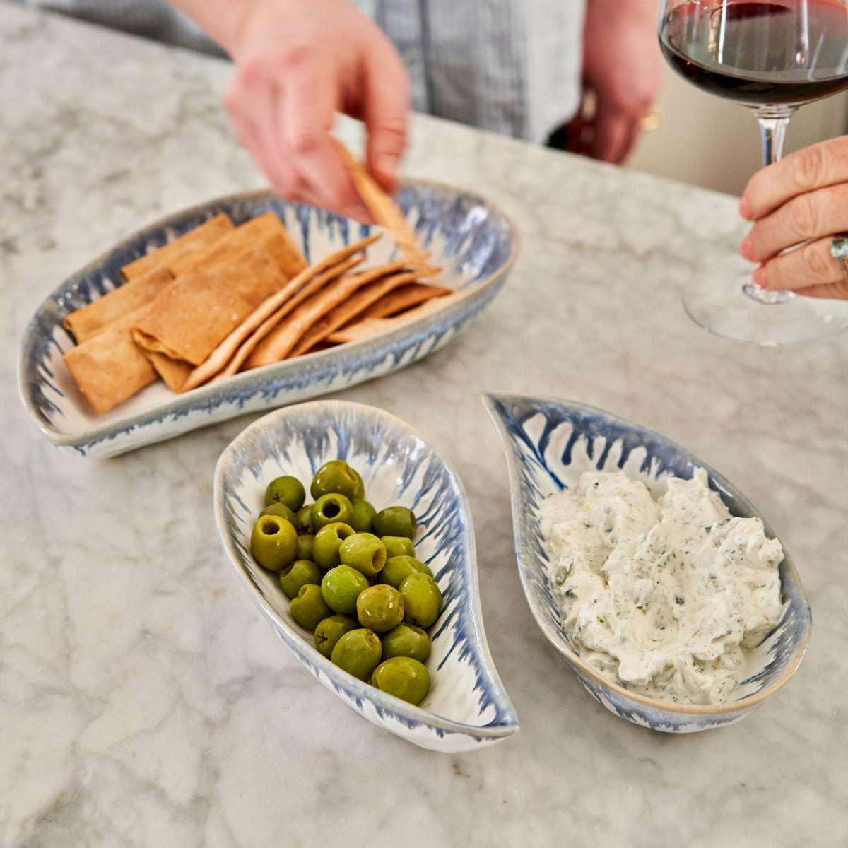 Three shell-shaped bowls in blue and white with olives, cheese dip, and crackers on a marble table.