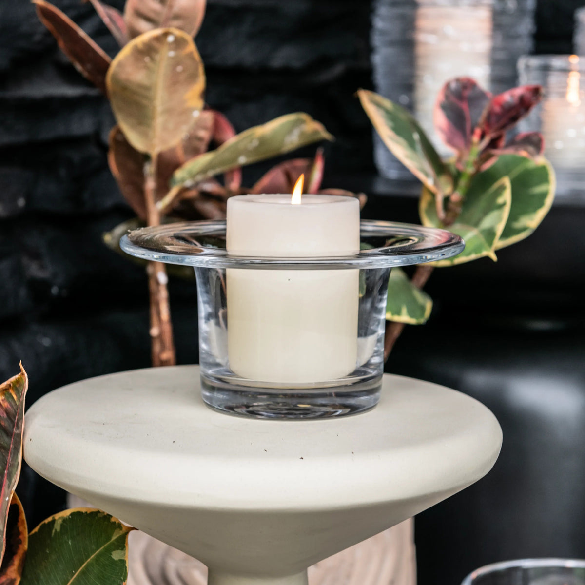 White pillar candle in clear glass holder with wide rim, positioned on cream pedestal base surrounded by potted plants.