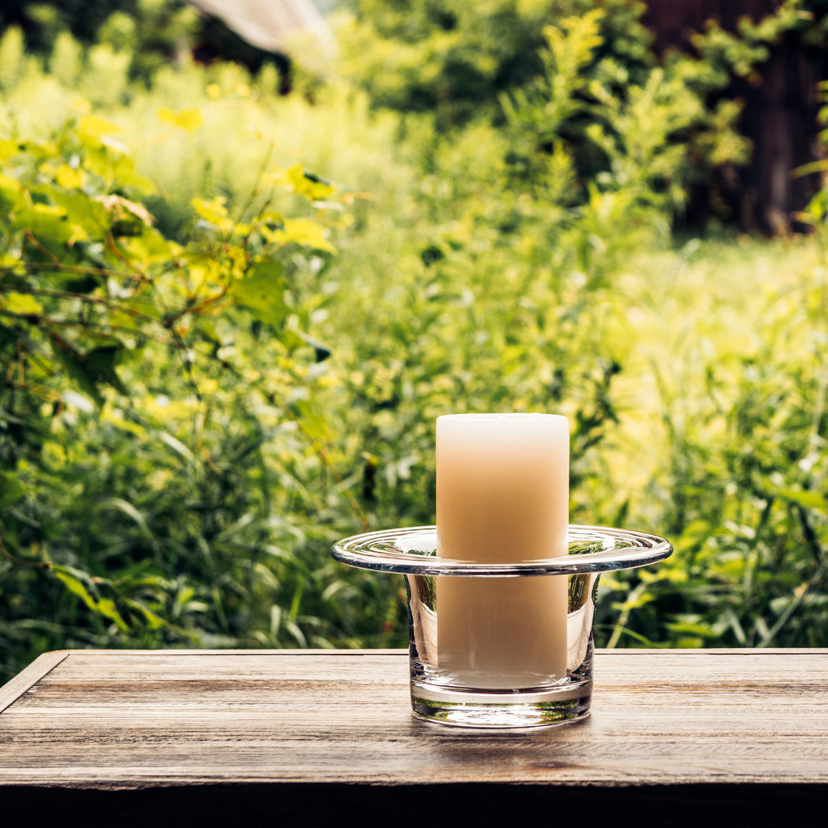 Cream-colored pillar candle in a clear glass hurricane holder on a wooden surface with green foliage background.